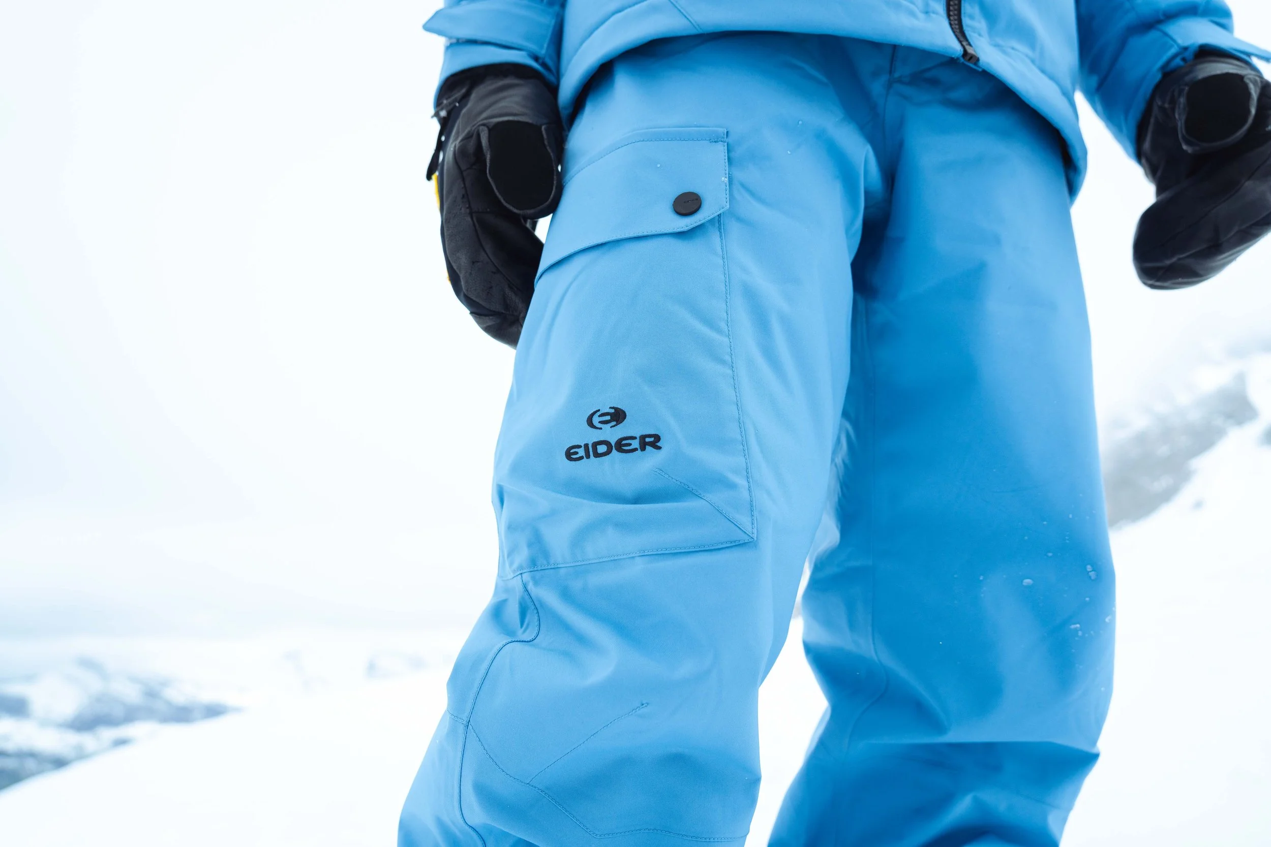 Vêtement de ski bleu Eider avec gants noirs en arrière-plan neigeux.