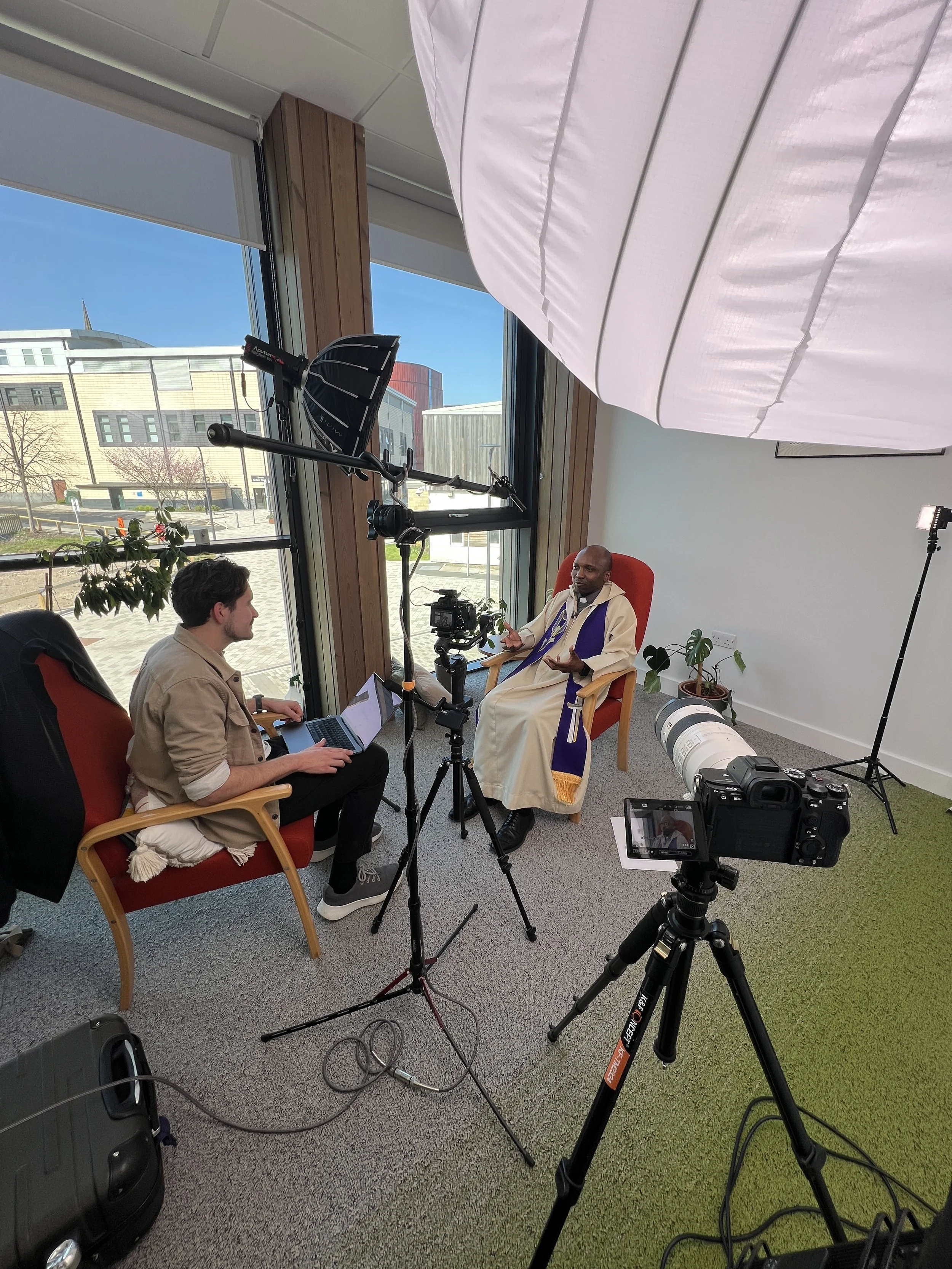 two men sat in red chairs next to a window, one holding laptop and other wearing chaplain outfit and being filmed by camera