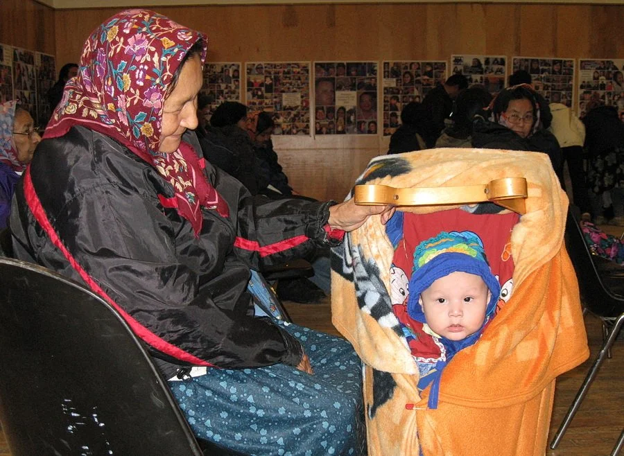 A First Nations Elder with her granddaughter in a tikanagan at an information meeting.