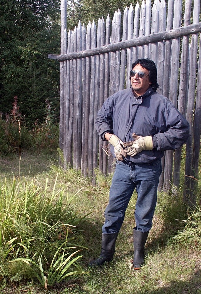 A First Nation man standing beside a palisade.