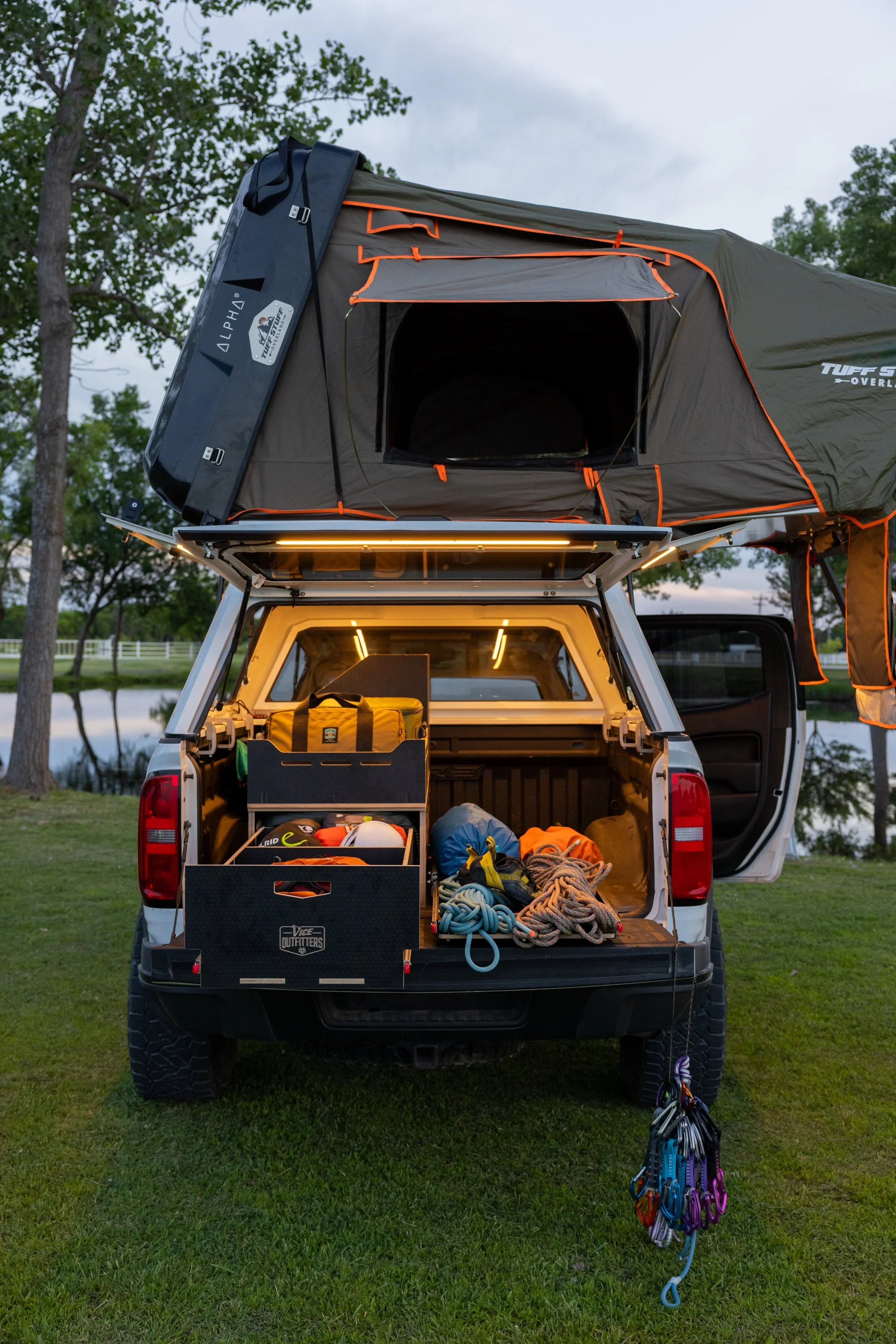 Back of a white truck with its tailgate down. A VICE Outfitters modular storage system packed with camping gear including ropes and storage boxes. A rooftop tent set up on top.