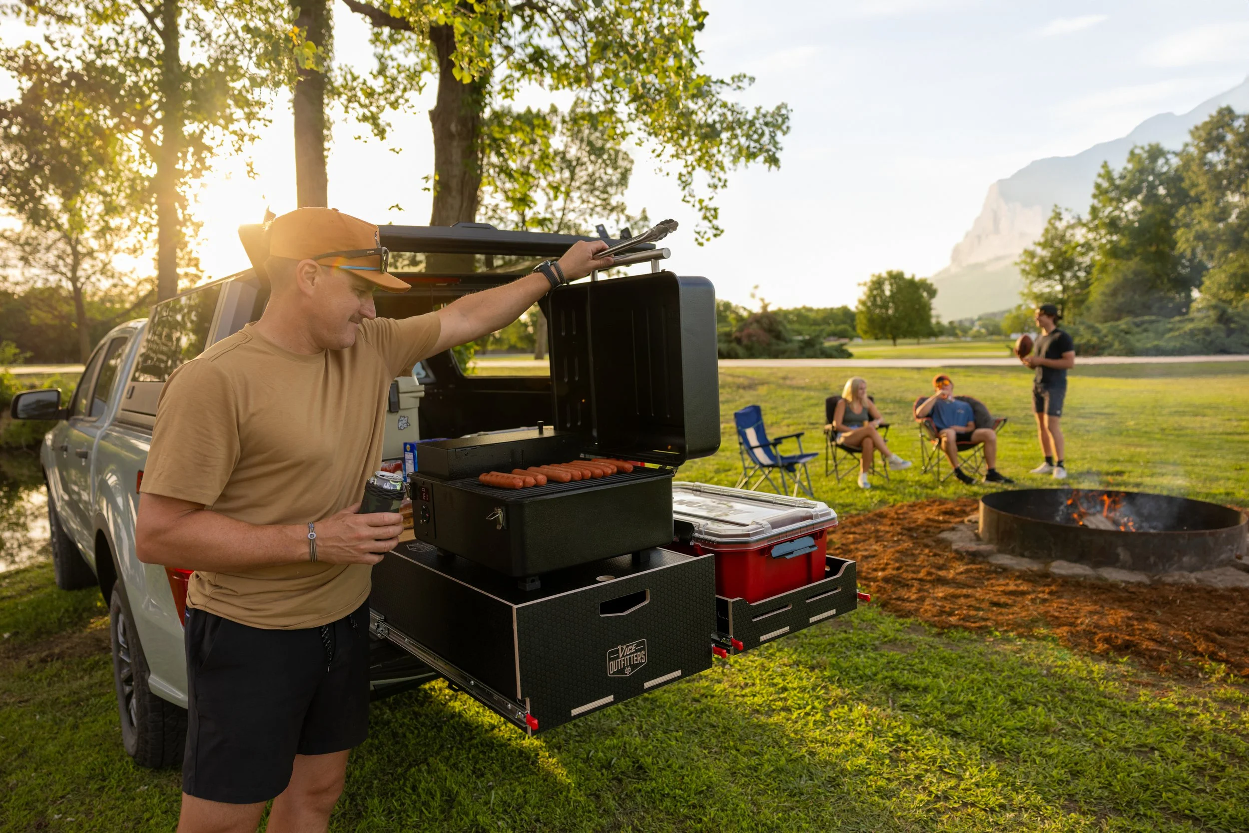 A man grilling hot dogs at a campsite next to a lake using a VICE Outfitters Canyon Drawer and Canyon Drawer Lid.
