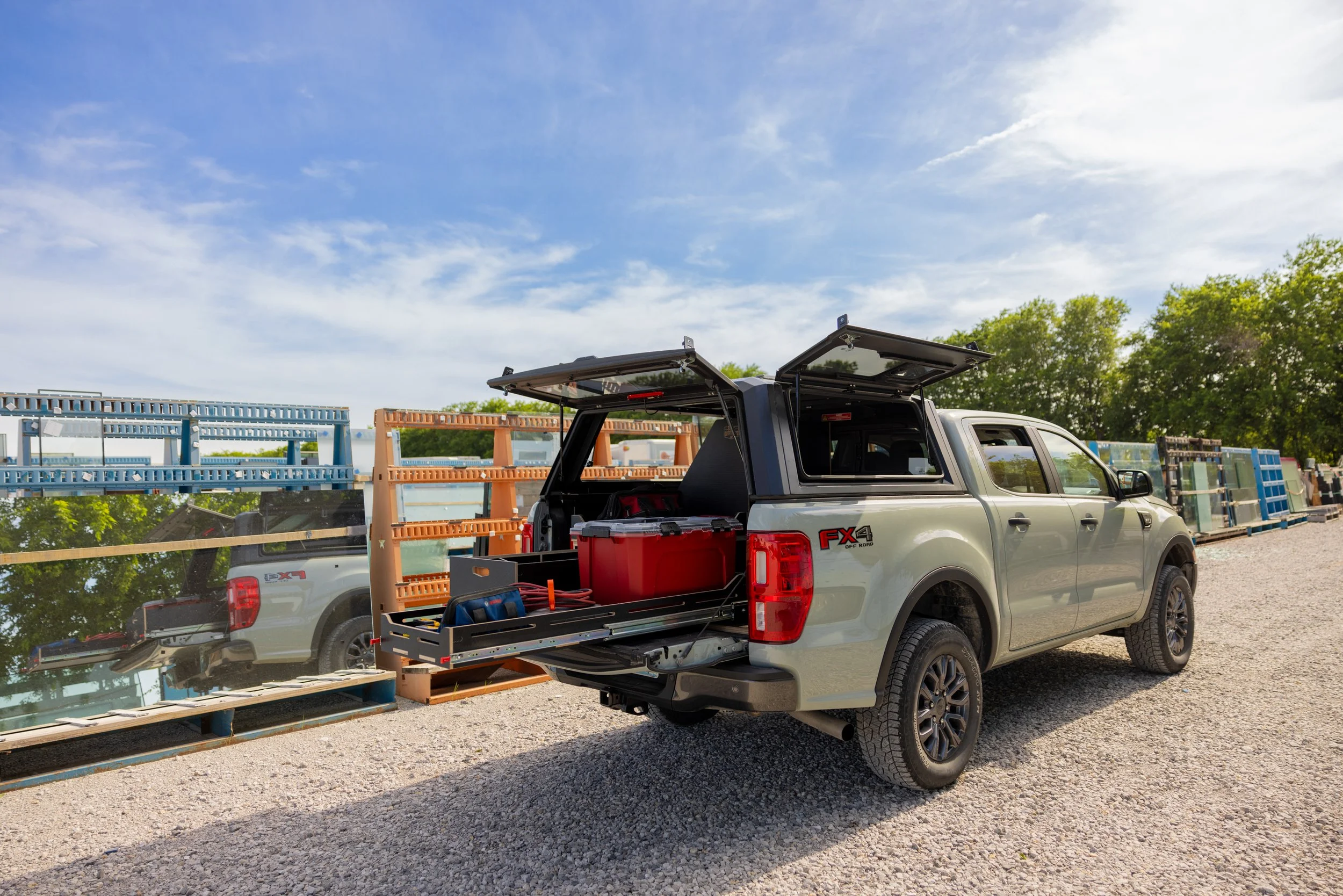 A gray pickup truck with open tailgate and overhead canopy, parked on gravel, with a VICE Outfitters modular storage system in the truck bed. There are industrial shelves and green trees in the background.