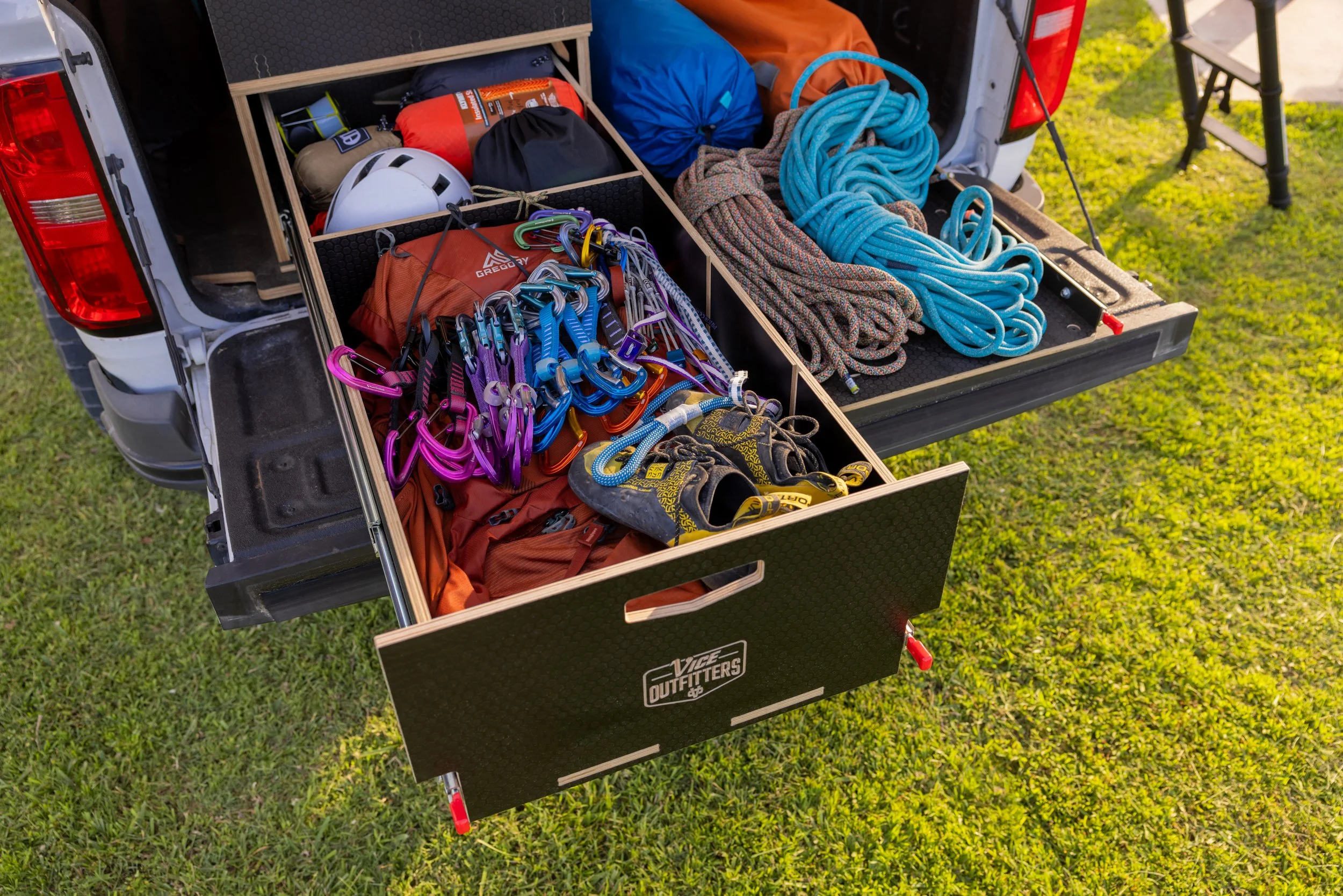Climbing gear organized in a VICE Outfitters Canyon drawer, including harnesses, ropes, helmets, and carabiners outdoors on grass.
