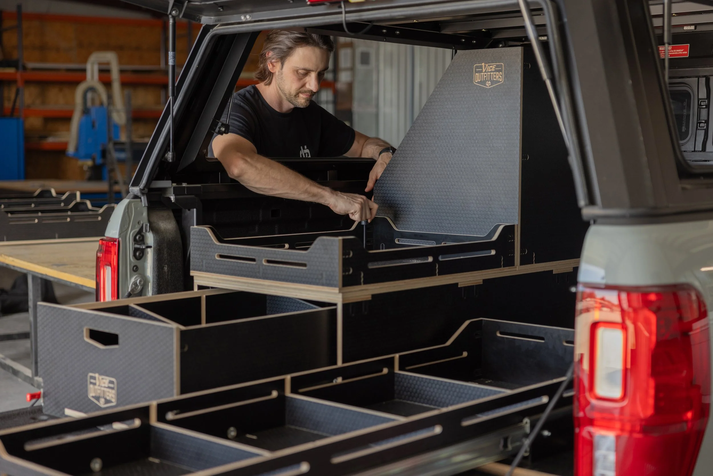 A man assembling a VICE Outfitters storage system in the back of a pickup truck in a workshop.