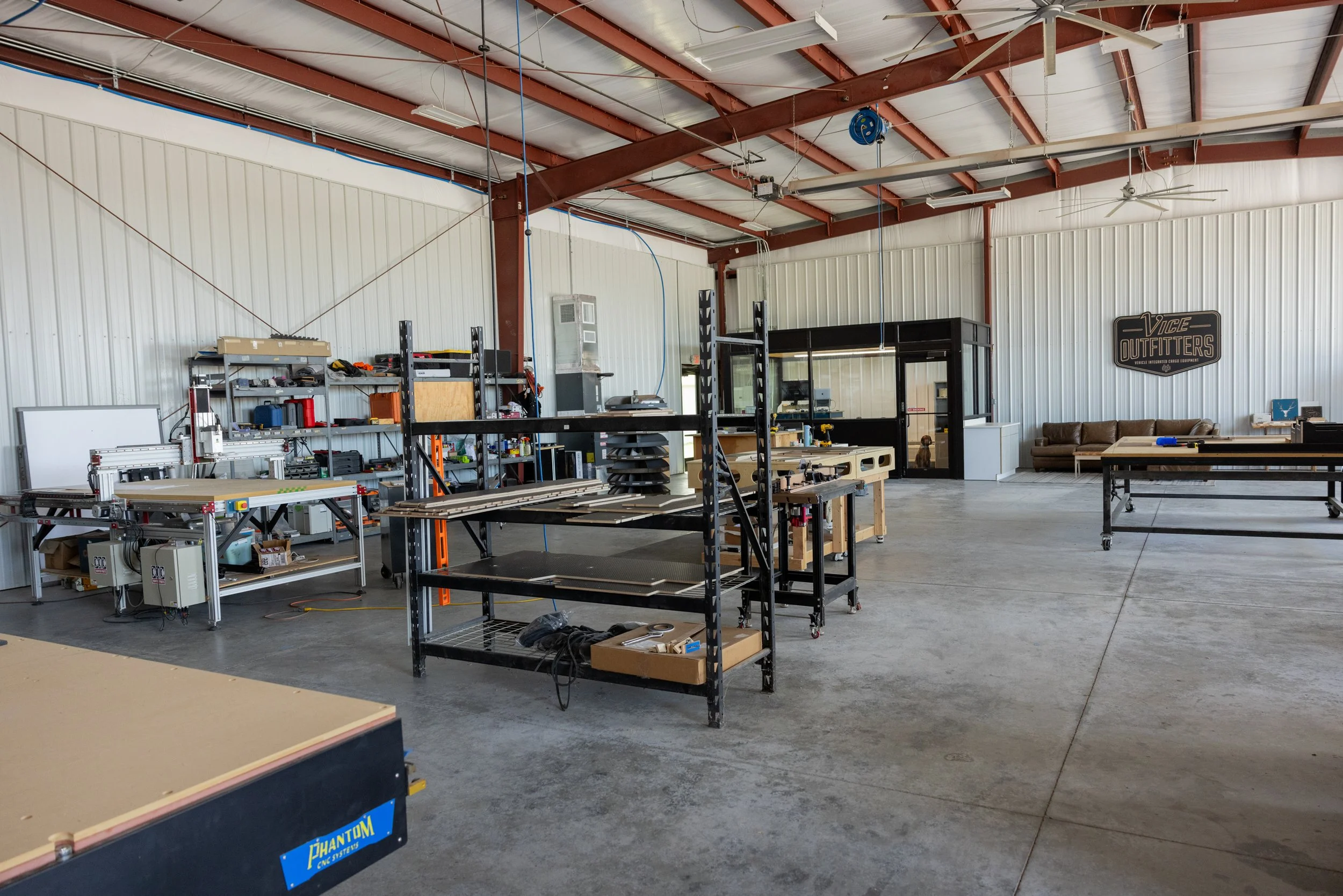 Interior of the VICE Outfitters shop with metal shelving, woodworking tools, and workbenches, including a CNC machine, in a spacious industrial-style building with a high ceiling and metal walls.
