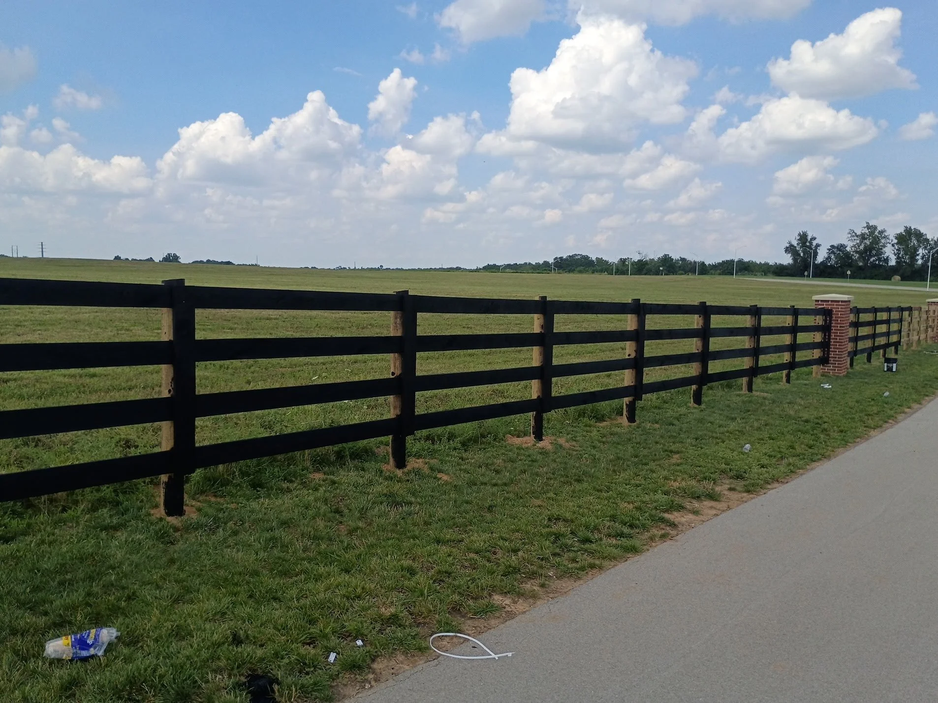 View of a countryside with a black wooden fence running parallel to a sidewalk, open grassy fields, a partly cloudy sky, a few trees in the distance, and some trash such as a plastic bottle and a white cable on the ground.