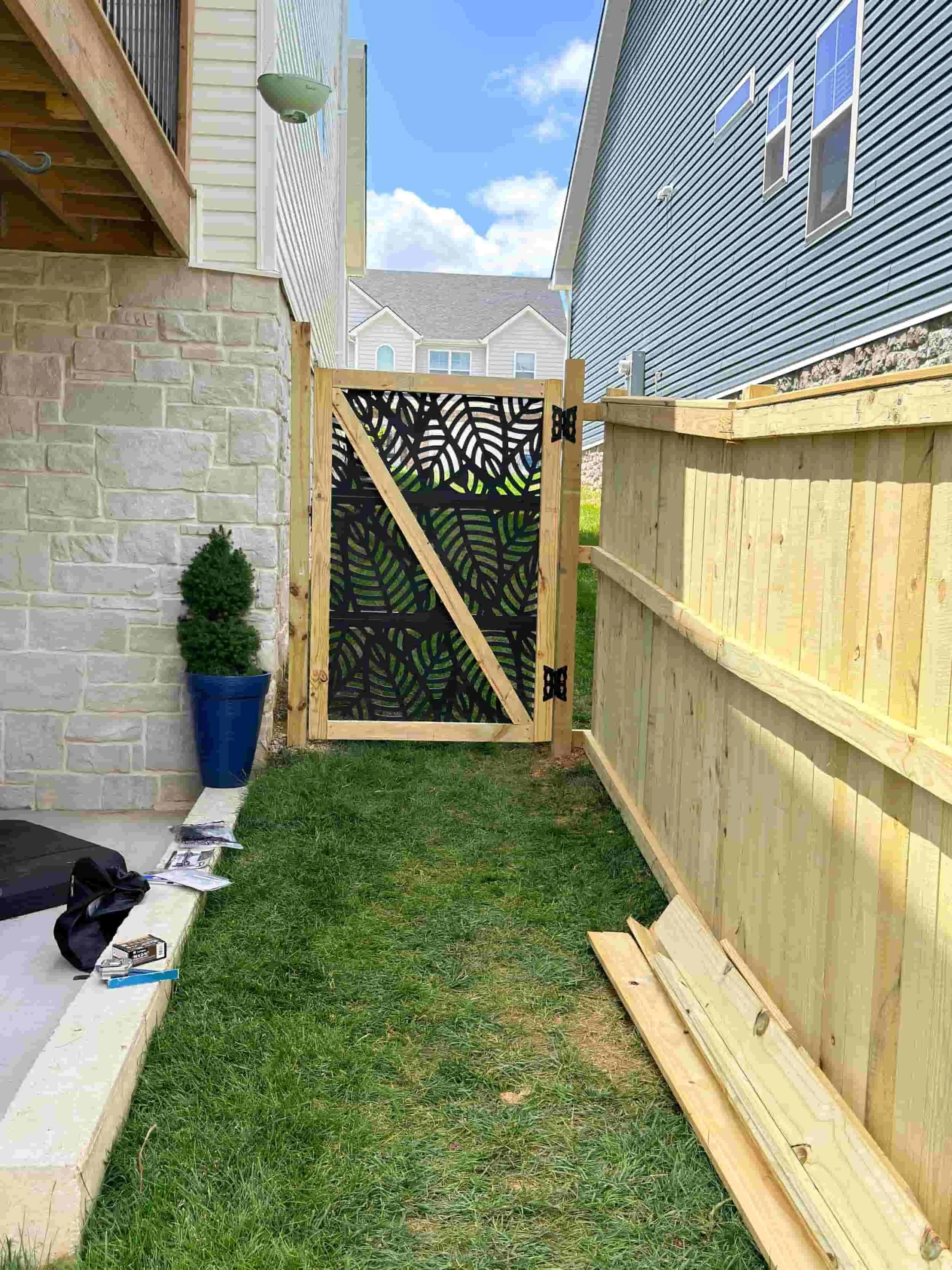 Side yard with wooden fence, partially built gate, small potted plant, tools, and construction materials on the grass under a blue sky with clouds.
