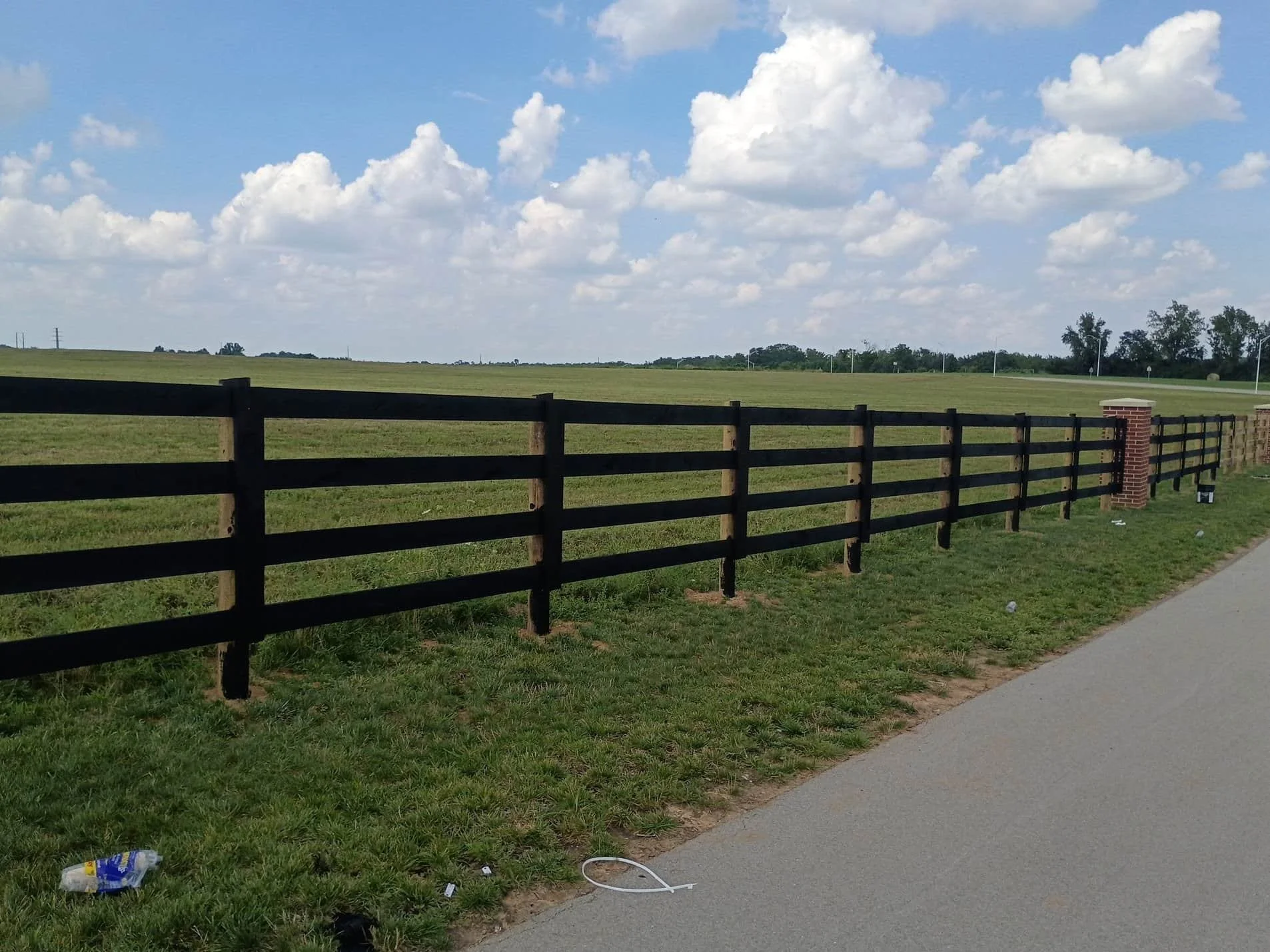 A black wooden fence runs along a sidewalk with green grass in front and a vast open field extending to the horizon. The sky above is mostly clear with scattered fluffy white clouds.