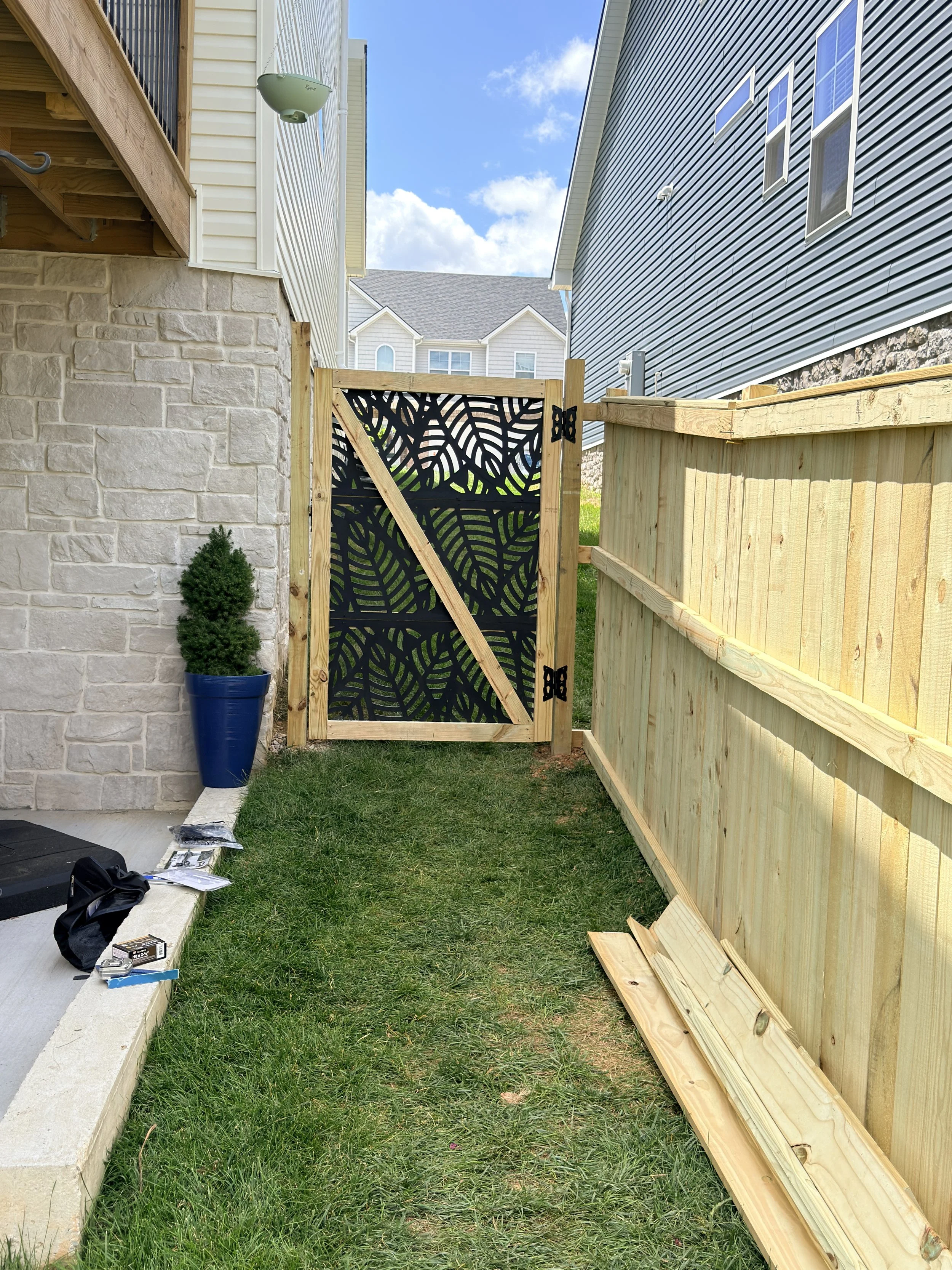 A backyard space under construction with a wooden gate, wooden fencing, a small potted shrub, building tools, and supplies on a concrete ledge, on a grassy area between two houses with siding.