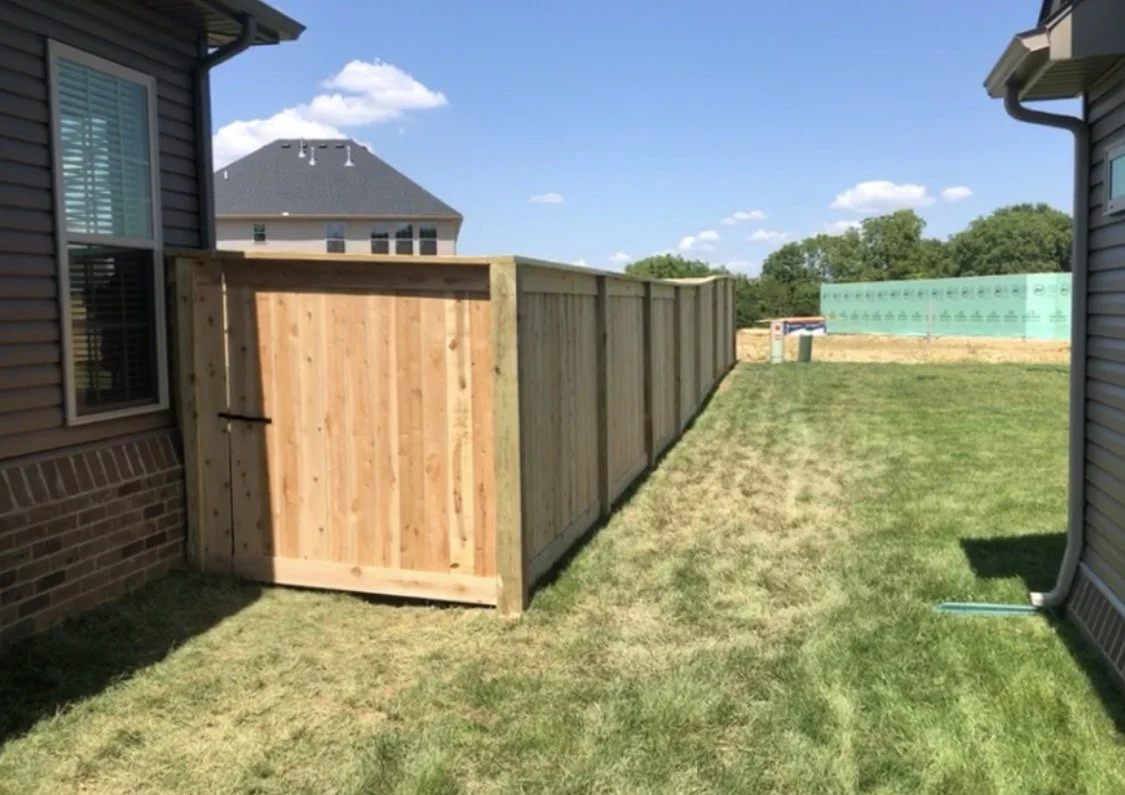 A backyard with a new wooden privacy fence between two houses, a grassy lawn, and a blue sky with a few clouds.