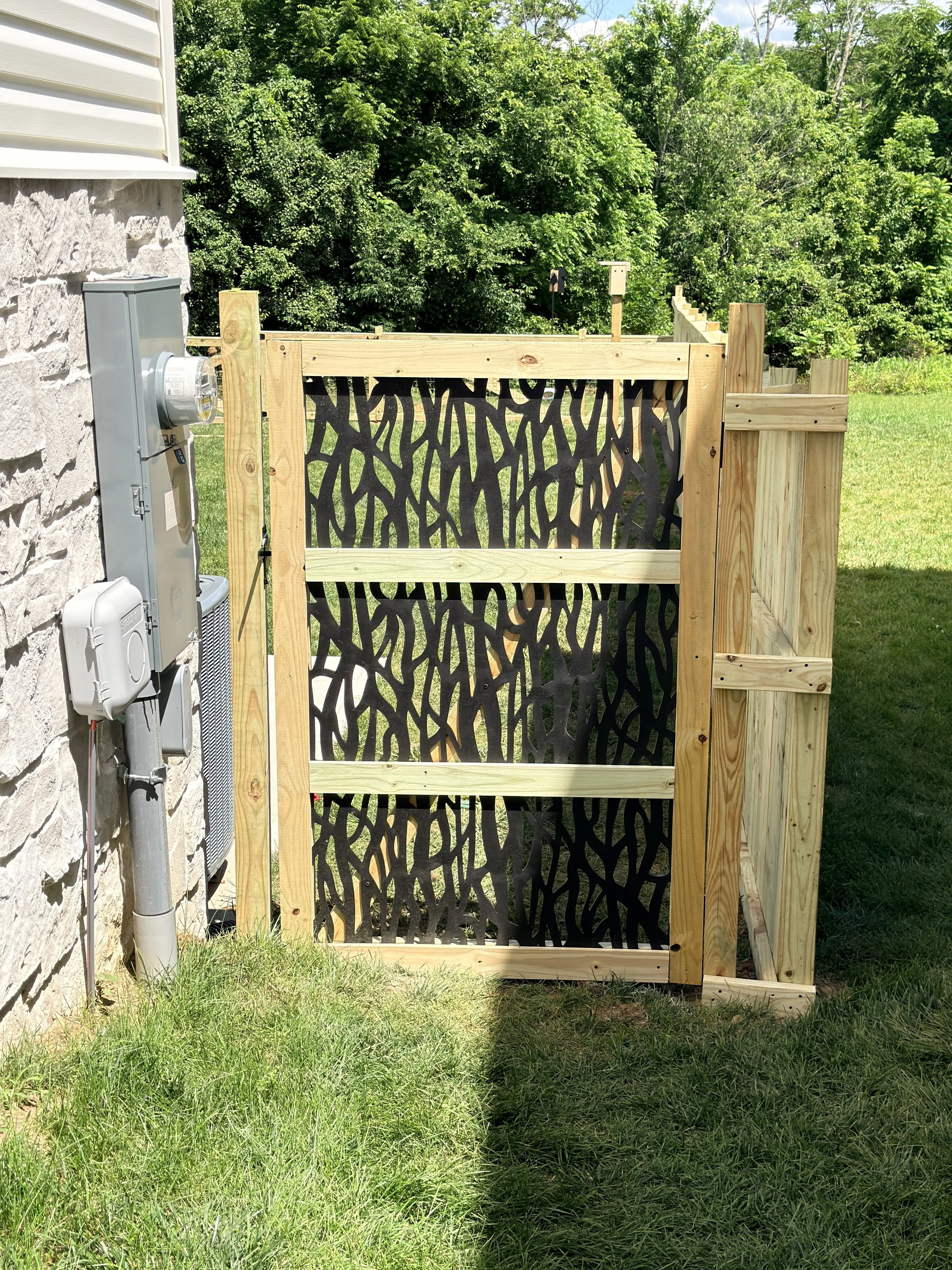 A wooden gate with black decorative metalwork installed outside of a house. To the left, installed on the house, is an electrical meter and box. The background features lush green trees and a grassy lawn.