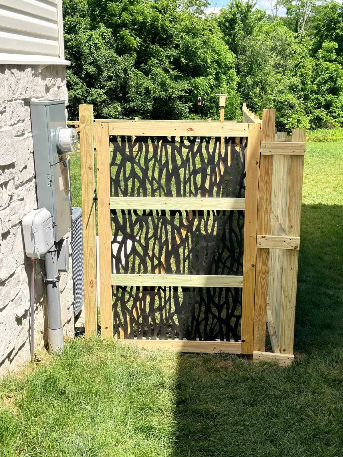Wooden gate with tree branch pattern installed next to a house with a stone exterior, behind a grassy yard, with lush green trees in the background.