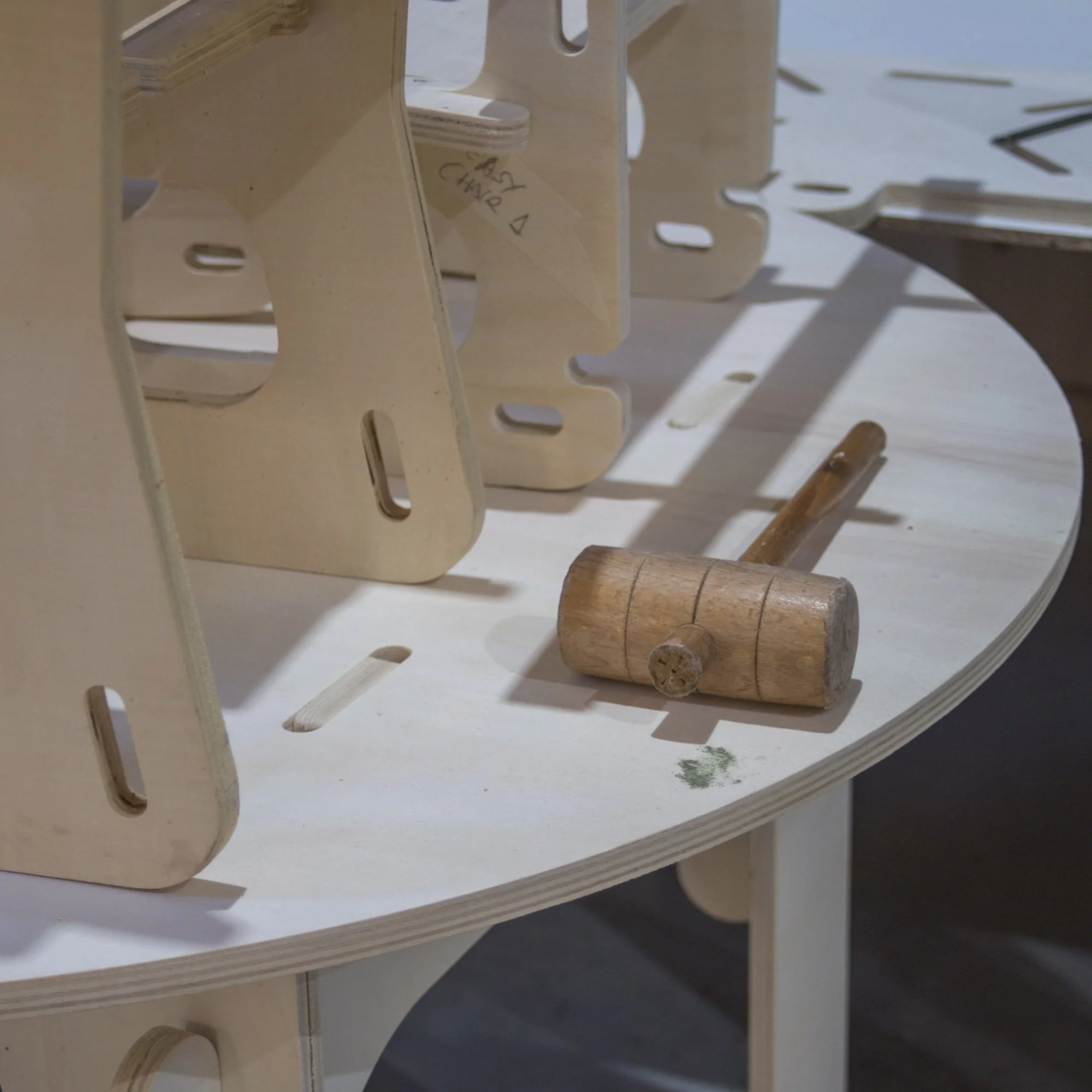 A wooden mallet lying on a white curved table, with assembled light-colored wooden pieces or structures surrounding it.