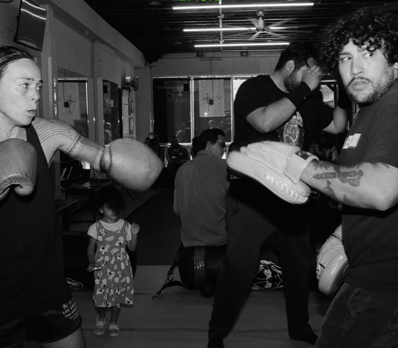 Two women sparring in boxing gloves in a boxing gym, with spectators and a young girl watching in the background, all in black and white.