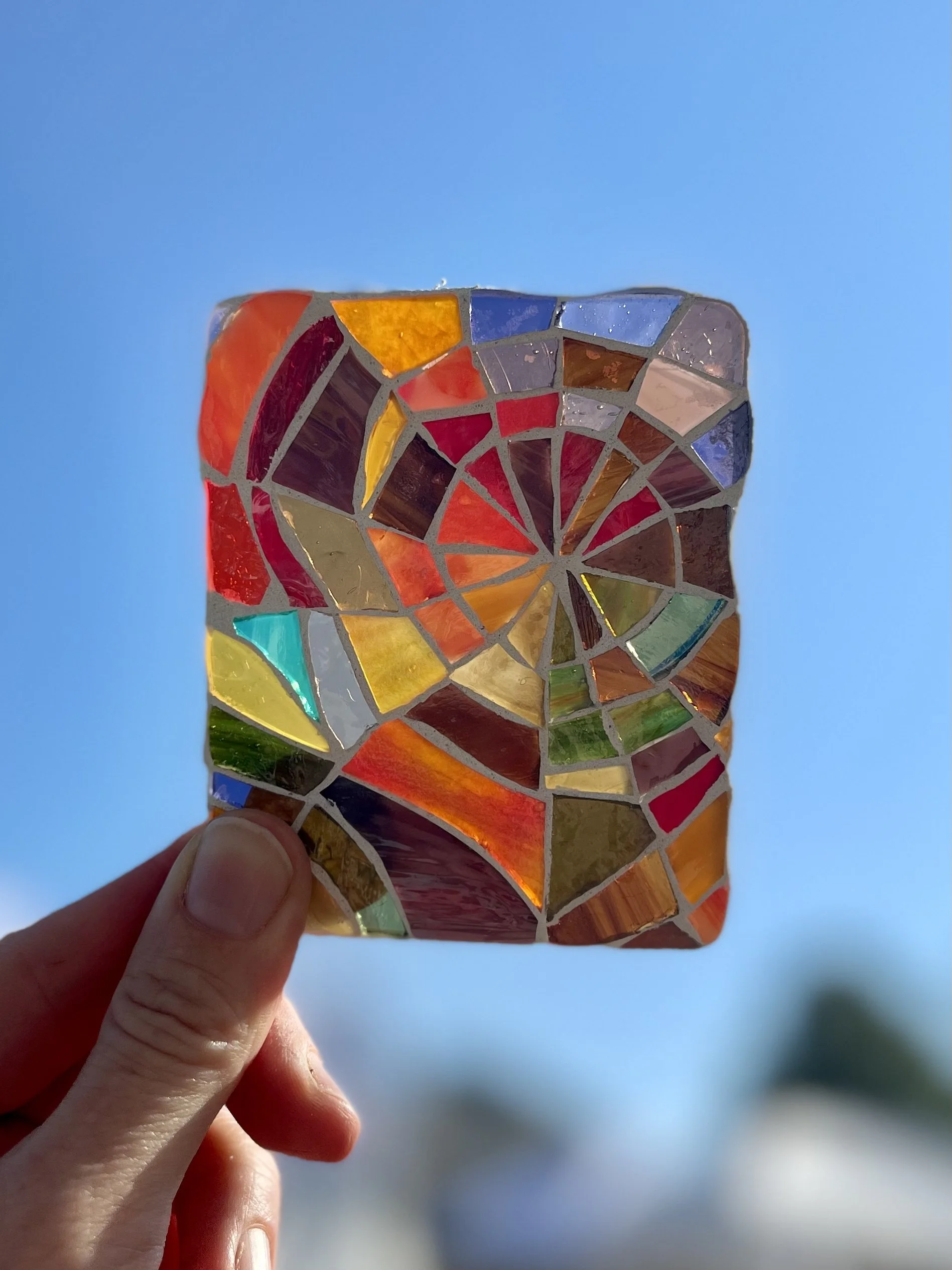 The artist's hand holding a colorful stained glass spiderweb mosaic rectangle against a blue sky.