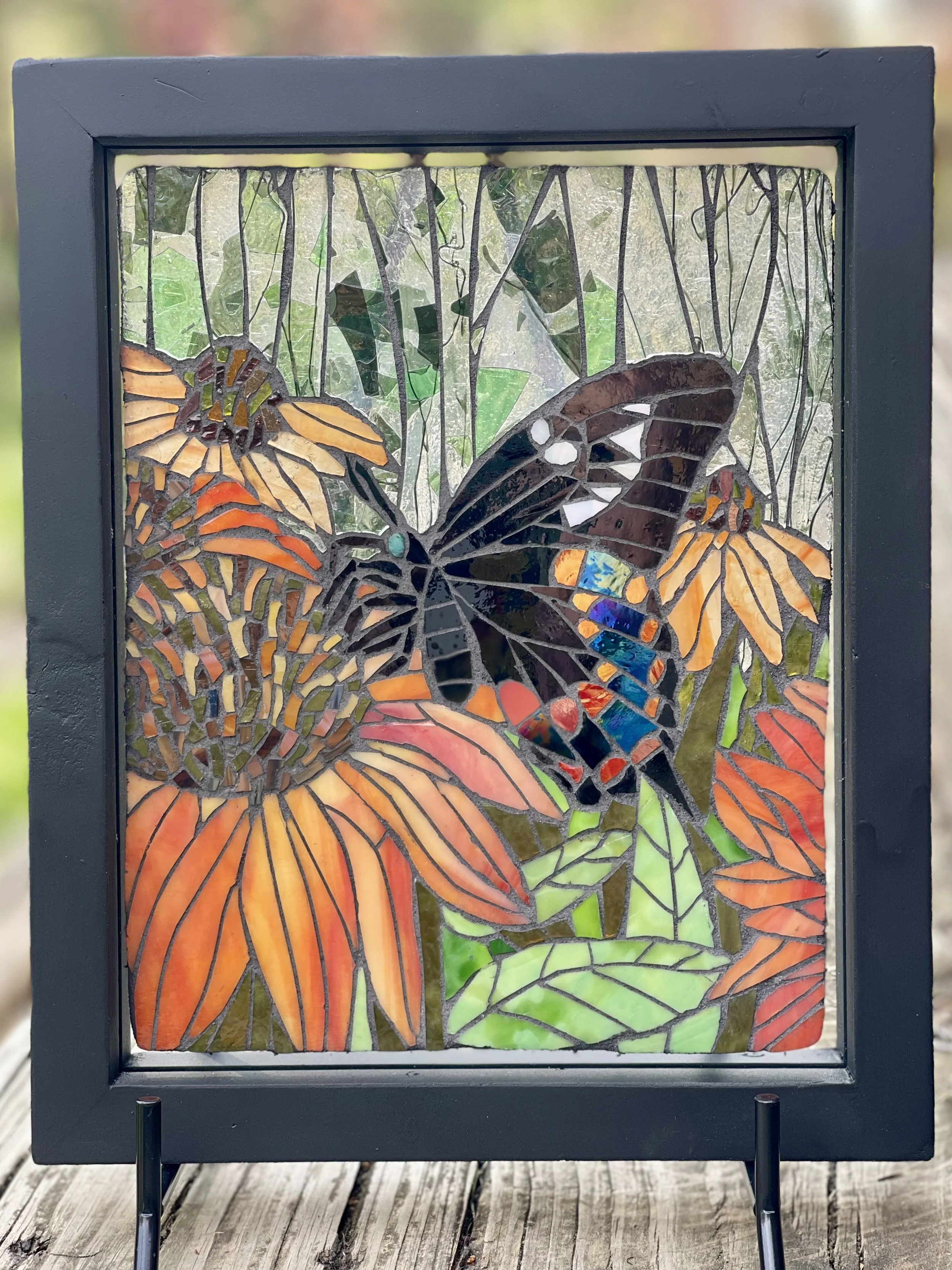 Stained glass artwork of a butterfly on orange and yellow flowers with green foliage background.