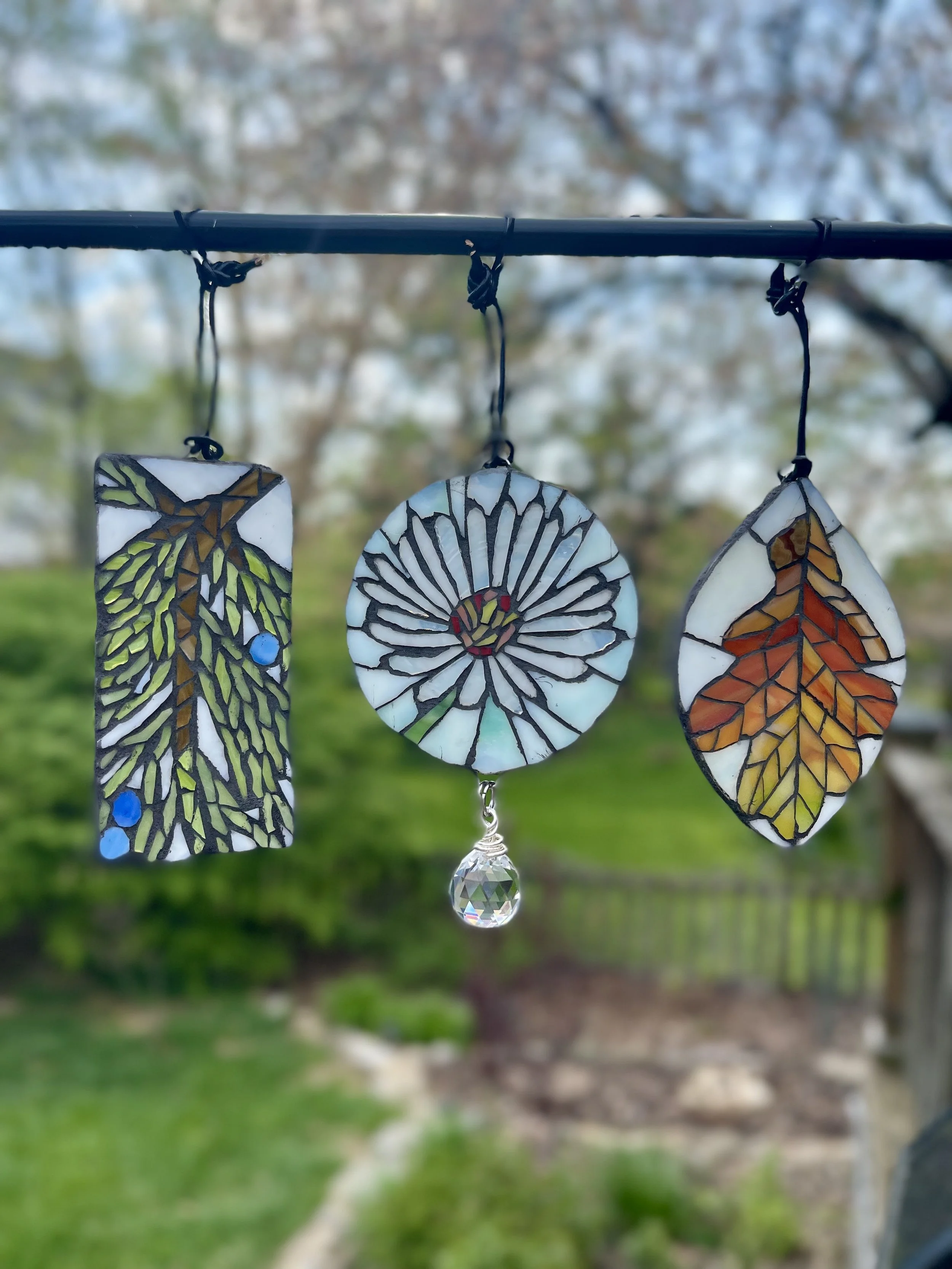 Three stained glass sun catchers (juniper with blue berries, a white aster, and an autumn oak leaf), hanging from a black rod outdoors on a cloudy day, with a background of trees and green grass.