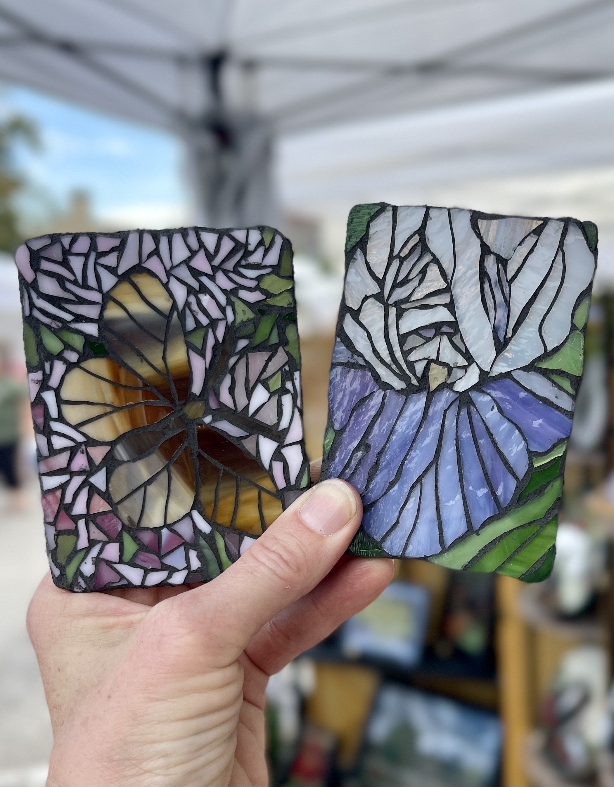 Two stained glass art pieces depicting a butterfly and a flower held up in the artist's hand, with a blurred outdoor market background.