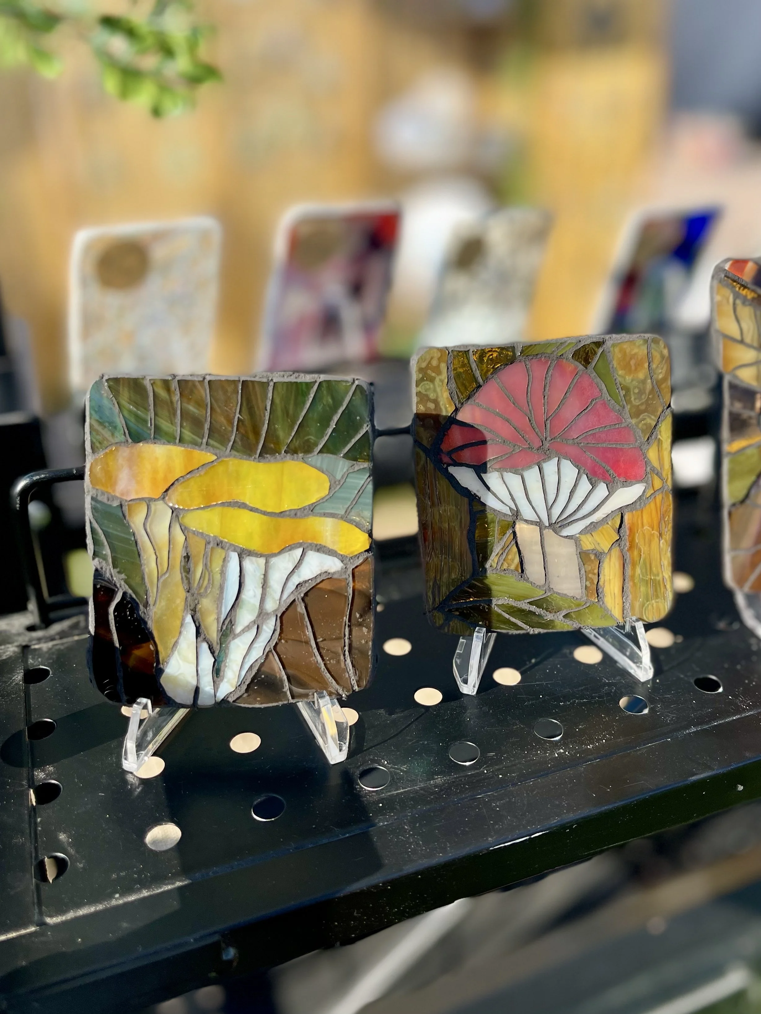 Colorful stained glass art pieces on display, featuring mushroom and plant designs, set on small stands outdoors.