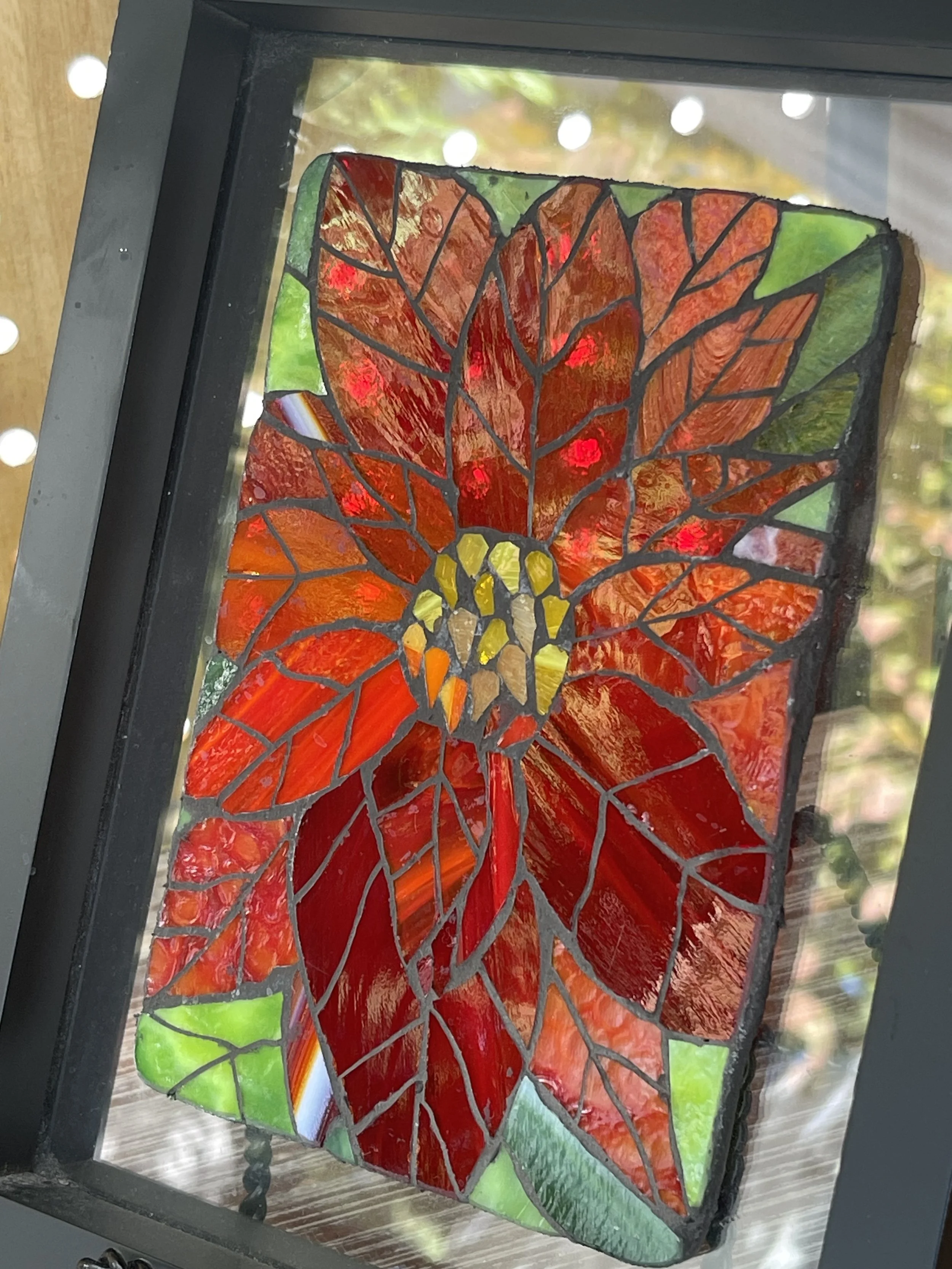 Colorful stained glass artwork of a Poinsettia with red, yellow, and green glass panels, framed in black.