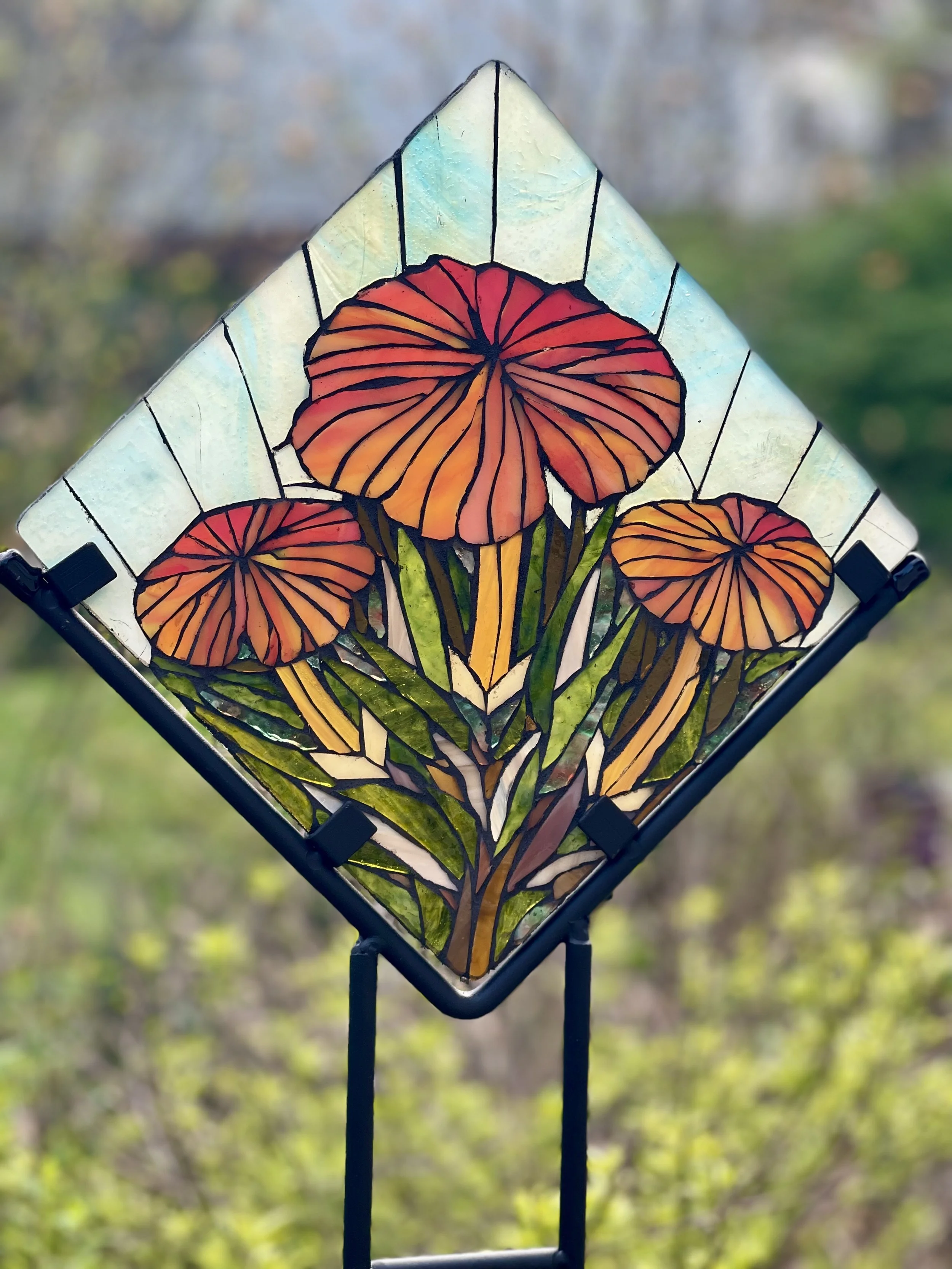 Colorful stained glass panel depicting three orange and red mushrooms with green stems and leaves, mounted on a black metal stand outdoors.
