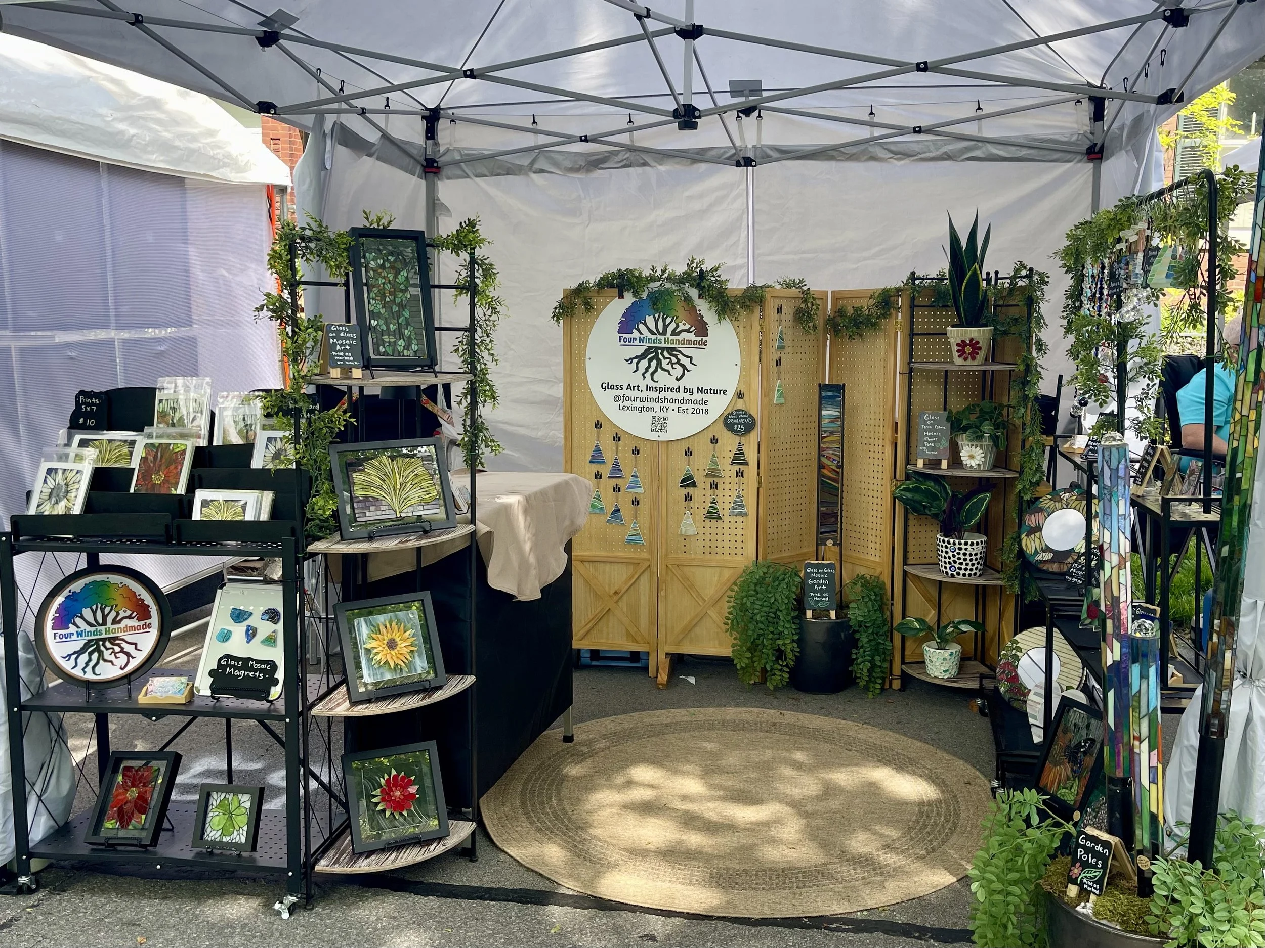 An outdoor art booth displays glass art inspired by nature, including framed stained glass art and hanging suncatchers, with greenery decoration and a circular rug on the ground.