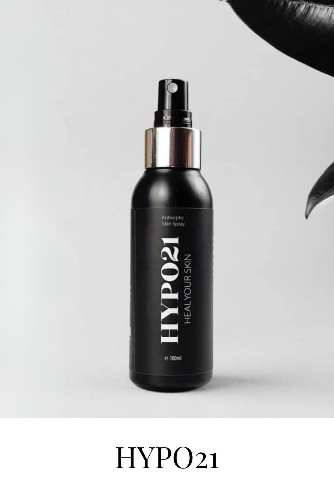 A black spray bottle labeled 'HYPO21 HEAL YOUR SKIN' with a silver cap and spray nozzle, standing against a plain light background, partially framed by a black leaf on the upper right and lower right corners.