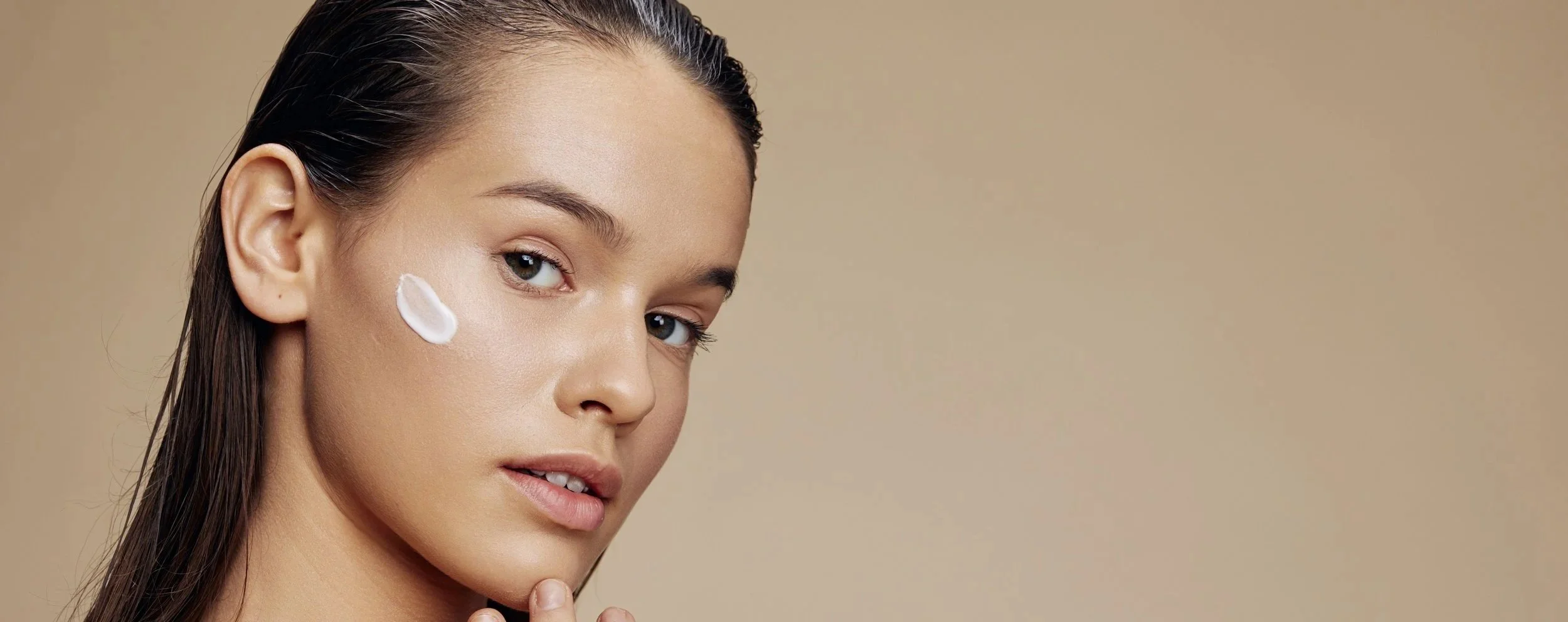 A woman with wet hair and light makeup, applying cream to her face, with a beige background.