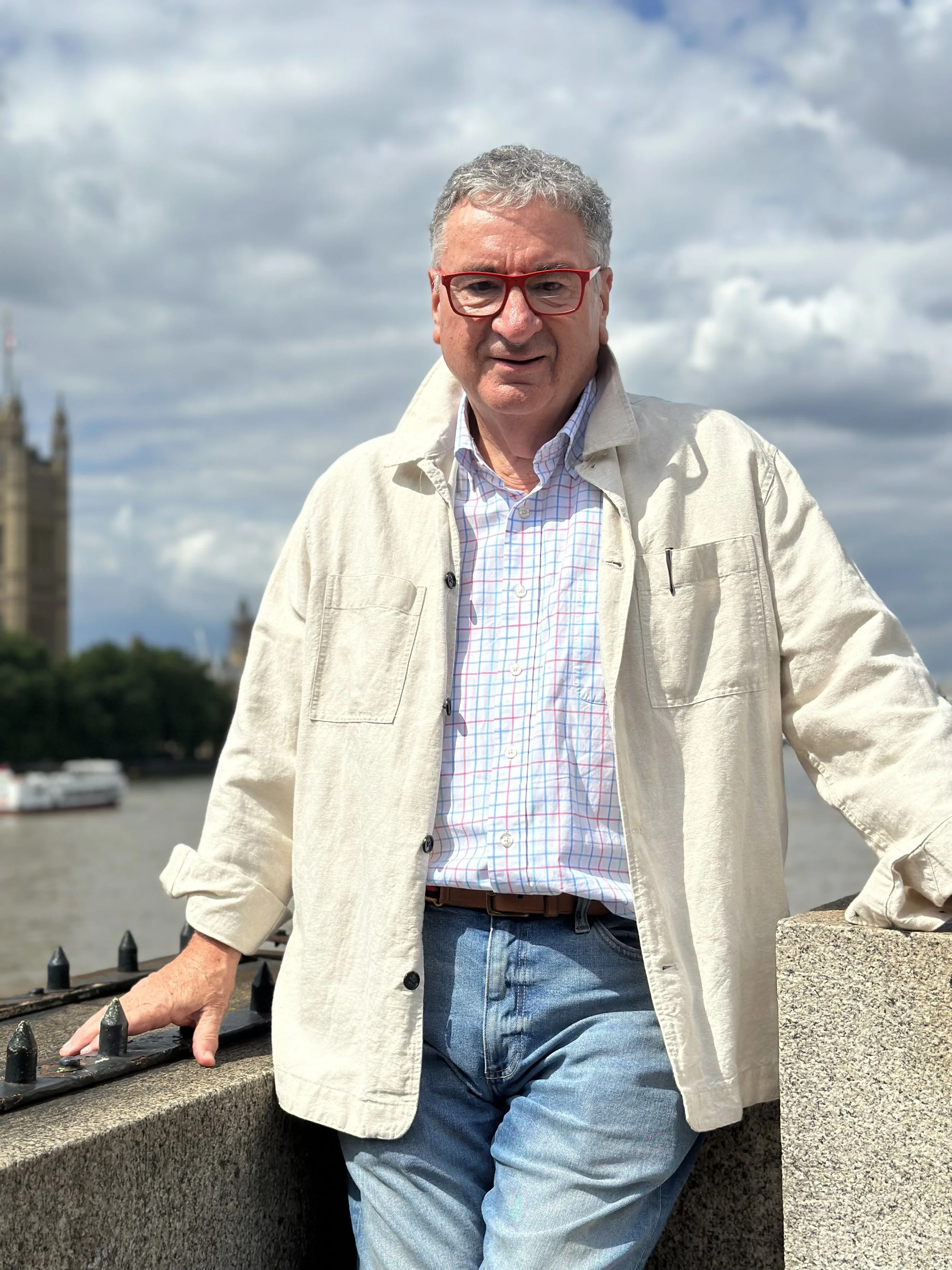 A man with gray curly hair and glasses standing by a river, with a cloudy sky and a building in the background.