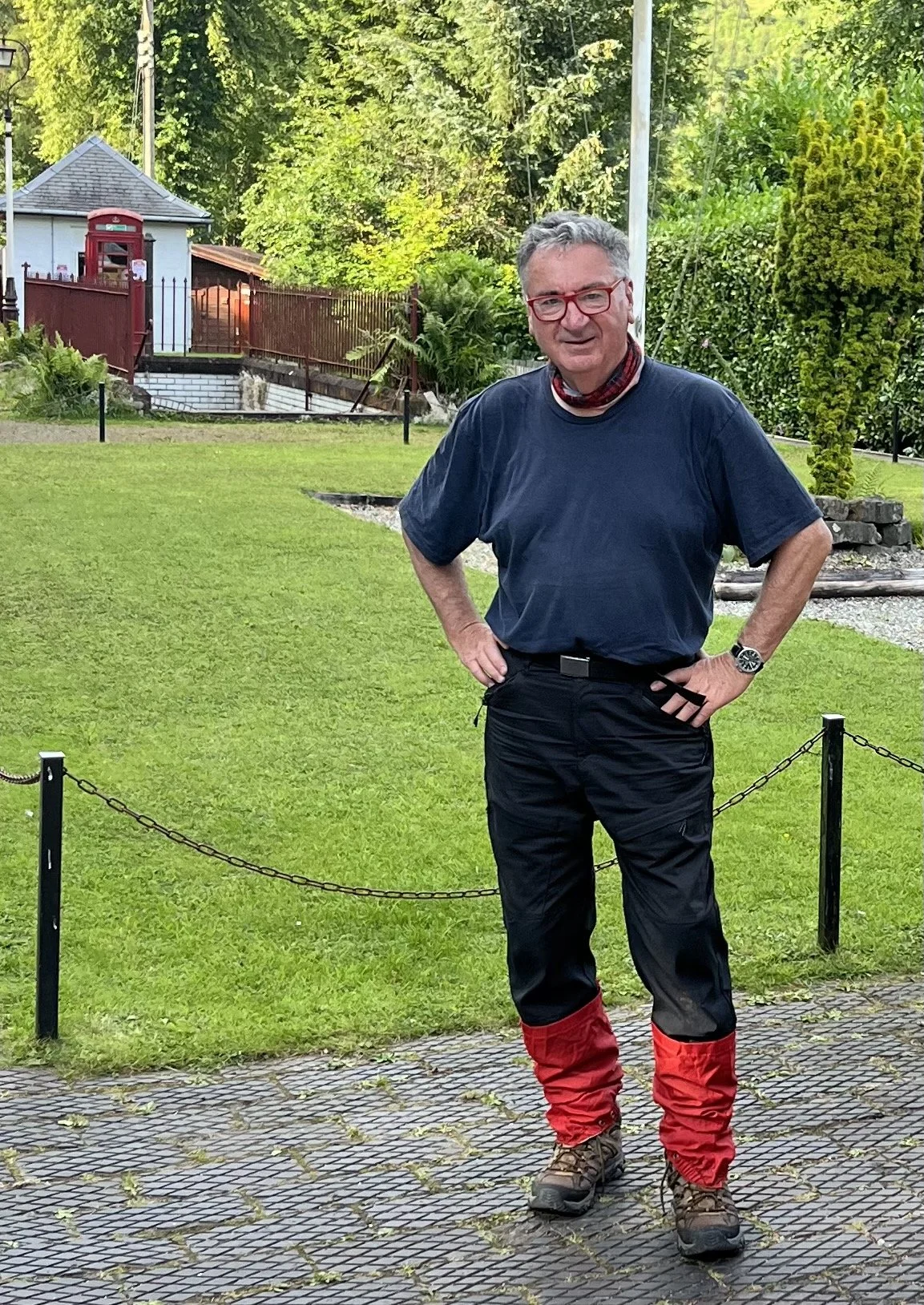 A man standing outdoors on a paved path, wearing outdoor gear and red gaiters, with a green lawn, trees, and a small building in the background.