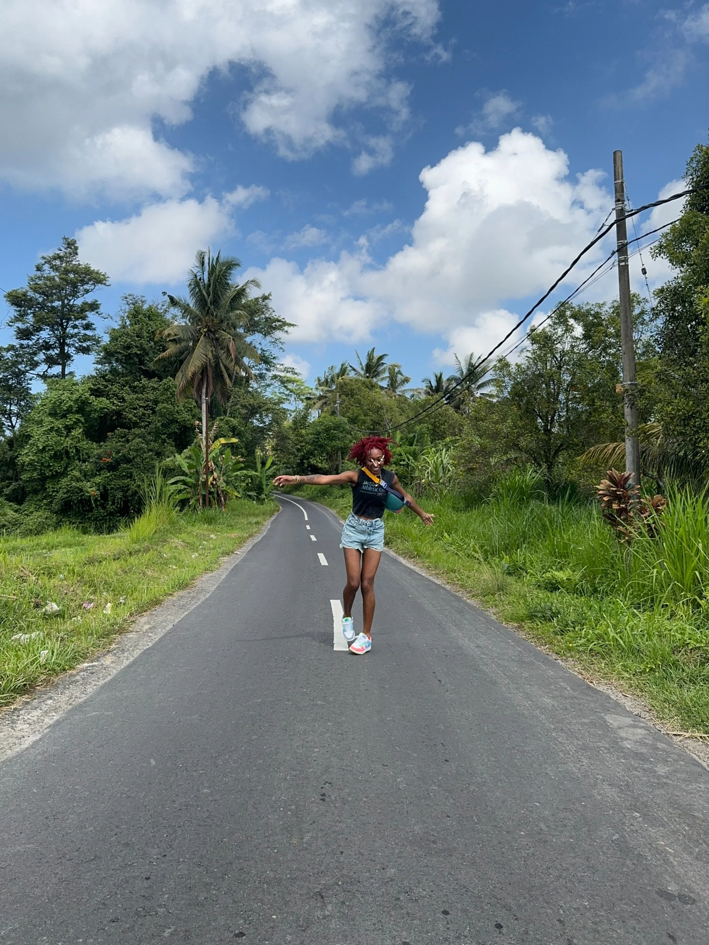 A woman with red hair, wearing a black shirt, denim shorts, and sneakers, balancing on a road surrounded by lush green vegetation and tall palm trees under a partly cloudy sky.