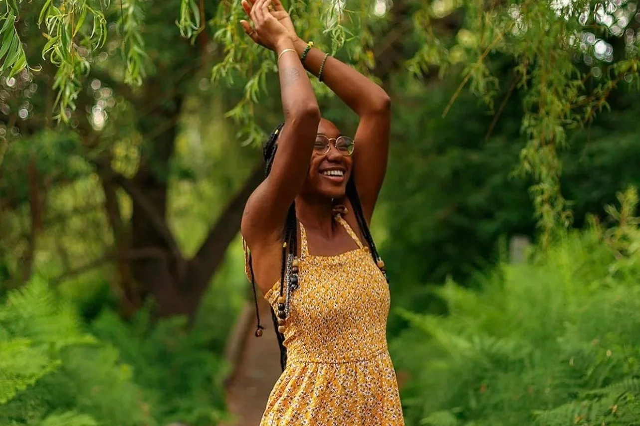A woman with braids and glasses smiling and stretching her arms upward among green trees and foliage.