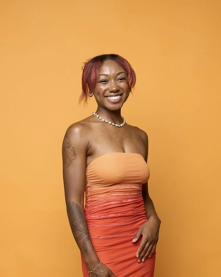 Young woman with red hair and tattoos, smiling, wearing a strapless orange to red gradient dress and pearl necklace, standing against a yellow background.