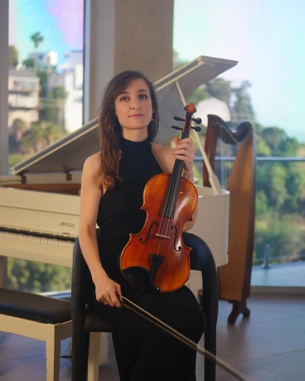 A woman in a black dress holding a violin and bow, sitting in front of a white grand piano inside a room with large windows showing trees and buildings outside.