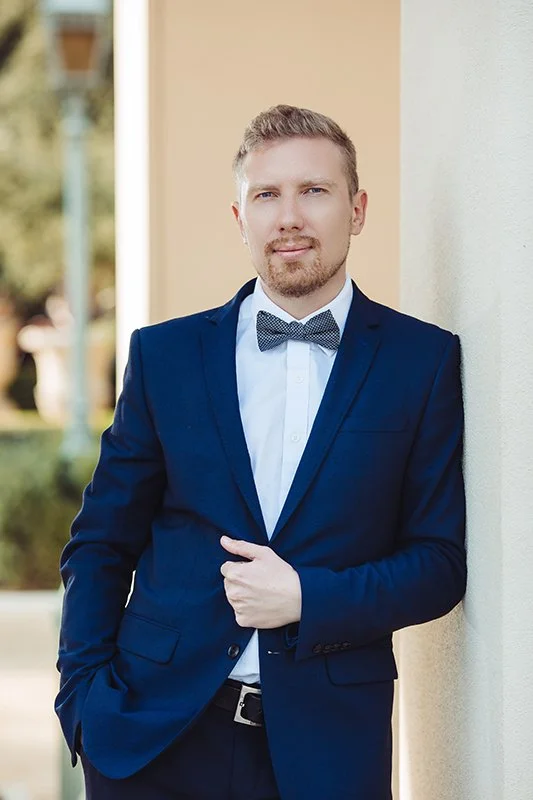 Man in a navy blue suit with a bow tie standing outdoors against a light-colored wall, with trees visible in the background.