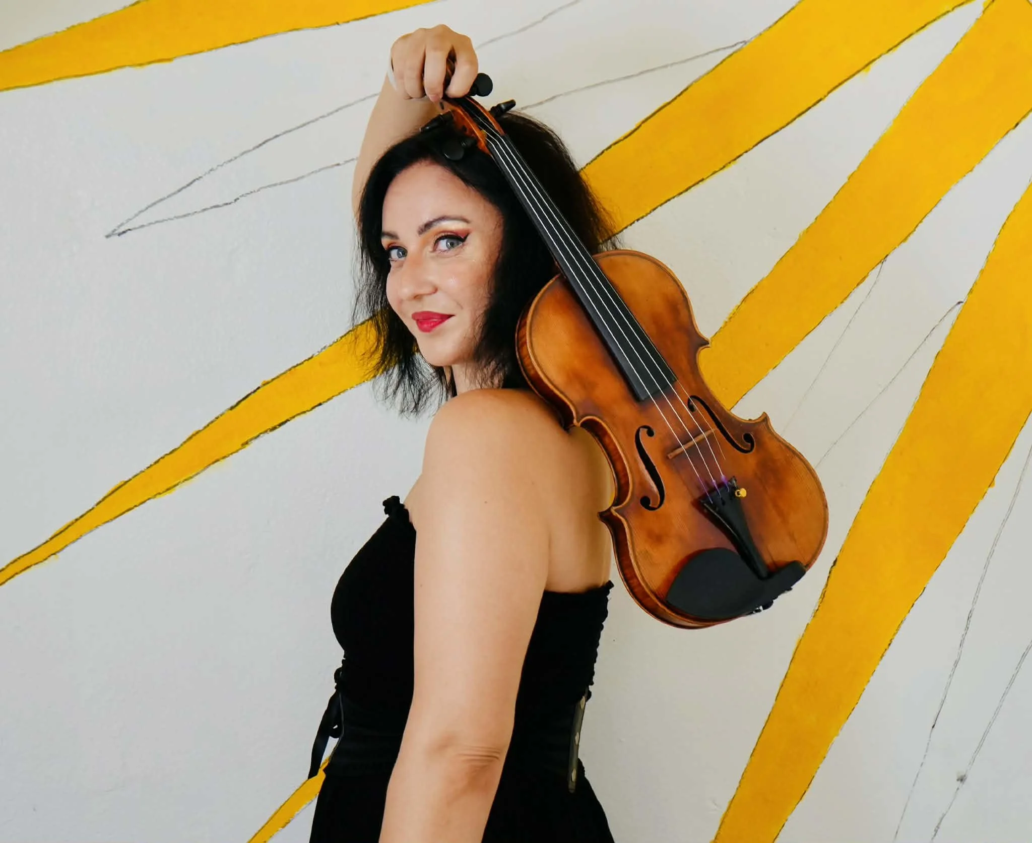 Woman with dark hair, red lipstick, and eye makeup holding a violin on her shoulder, standing in front of a white wall with yellow and black abstract lines.
