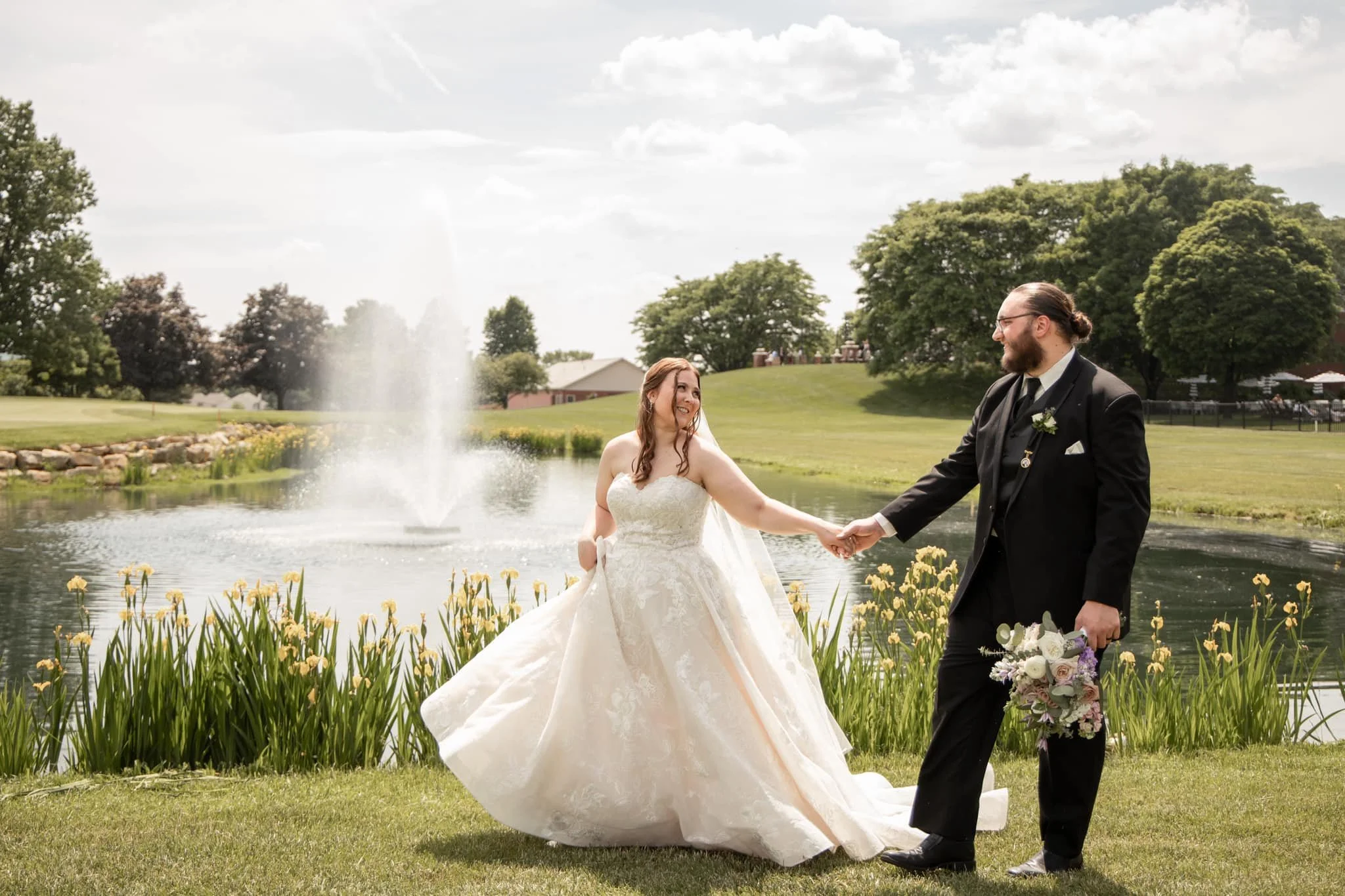 A bride and groom holding hands near a pond with a fountain, surrounded by grass and flowers, on a partly cloudy day.