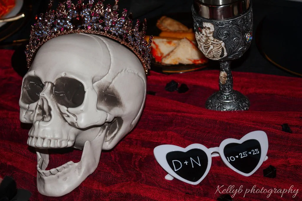 A white skull with a jeweled crown on its head on a red cloth table, next to a decorative goblet, black and transparent stones, a plate of sandwiches, and white heart-shaped sunglasses with black lenses showing the text "D+N" and the date "10.25.25".