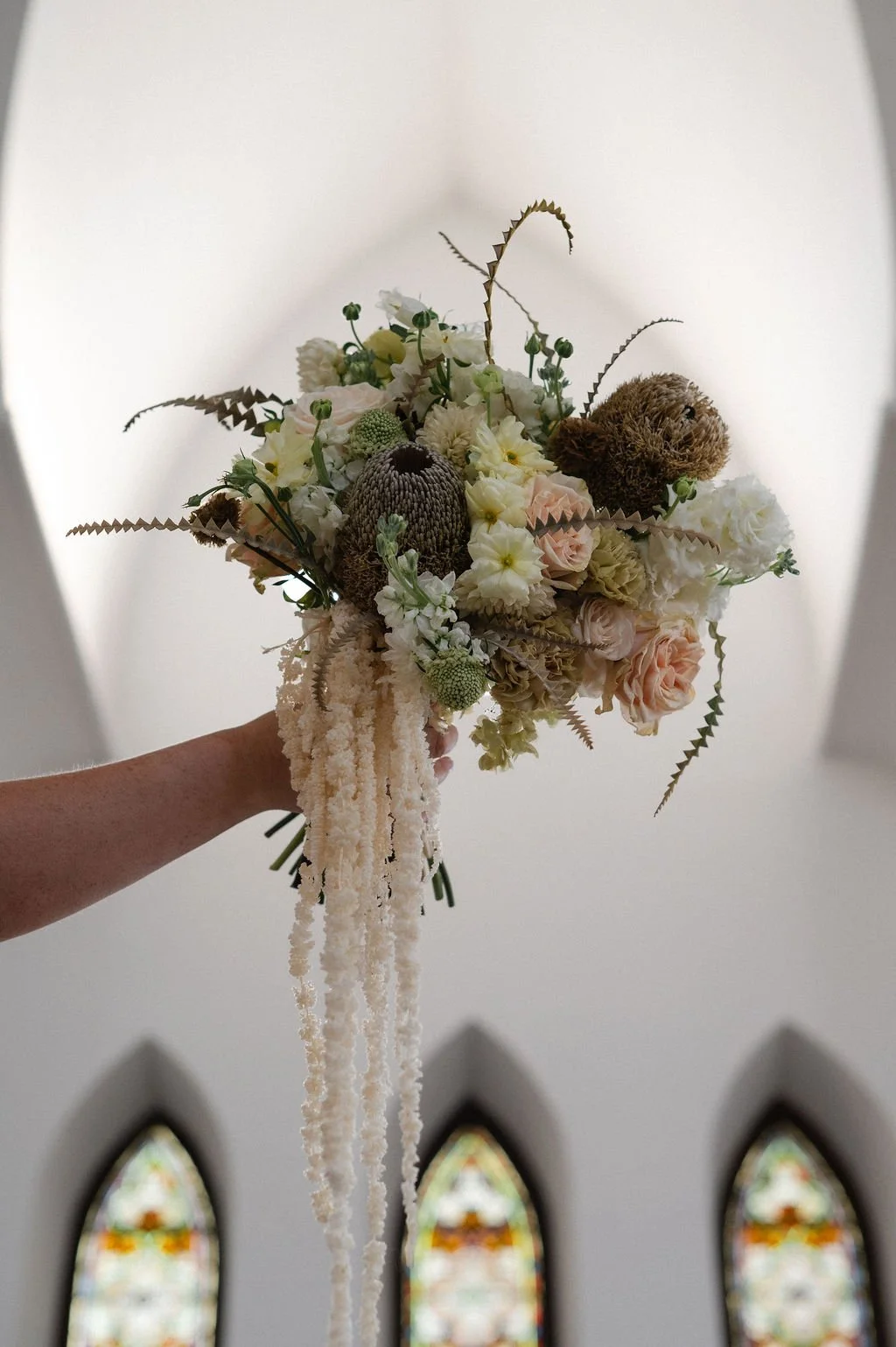 A hand holding a beige and white floral bouquet with various flowers and greenery, with stained glass windows in the background.