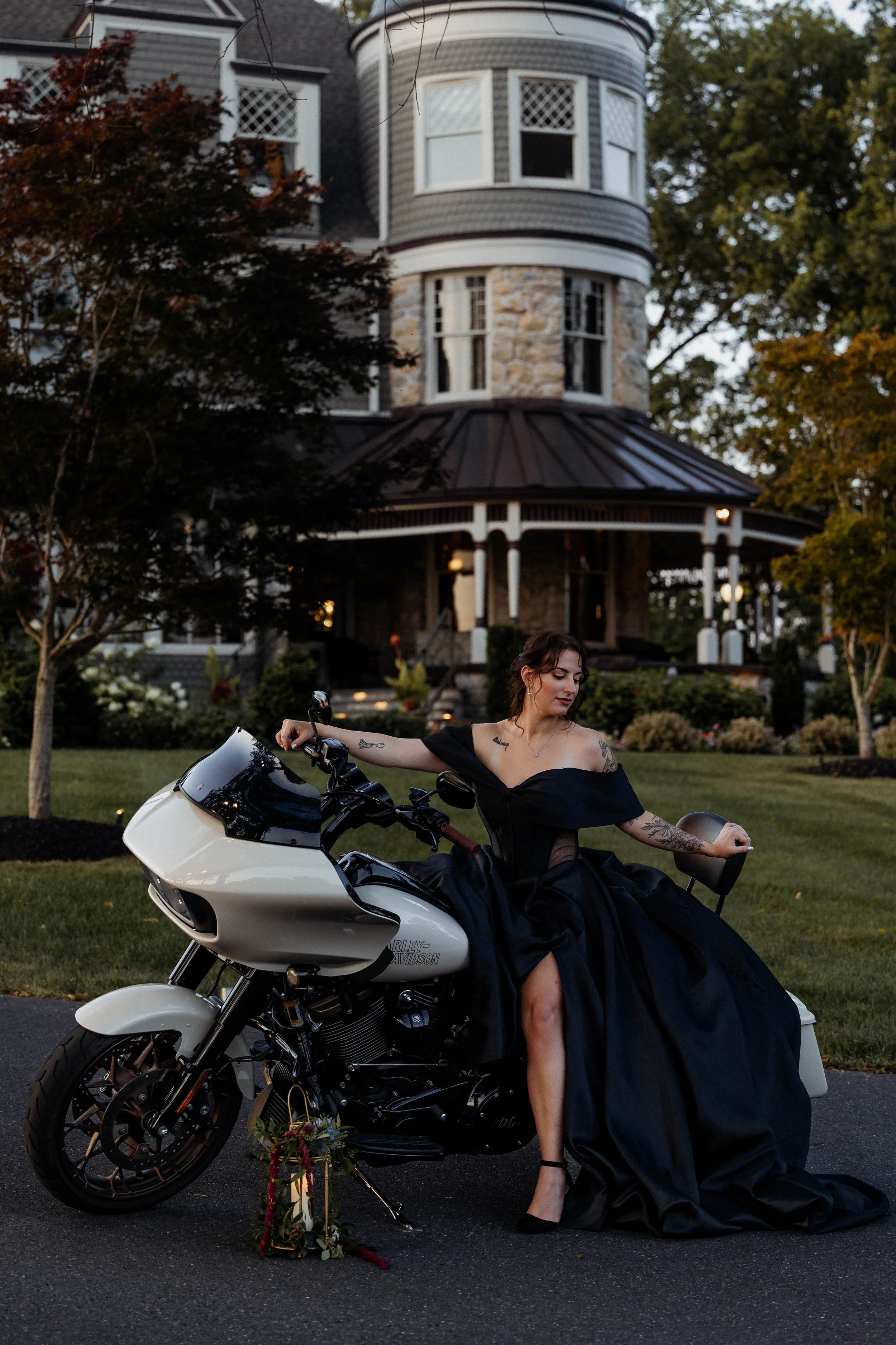 A woman in a black off-the-shoulder gown sitting on a white motorcycle in front of a large historic house with purple shingles and a front porch, during the evening.