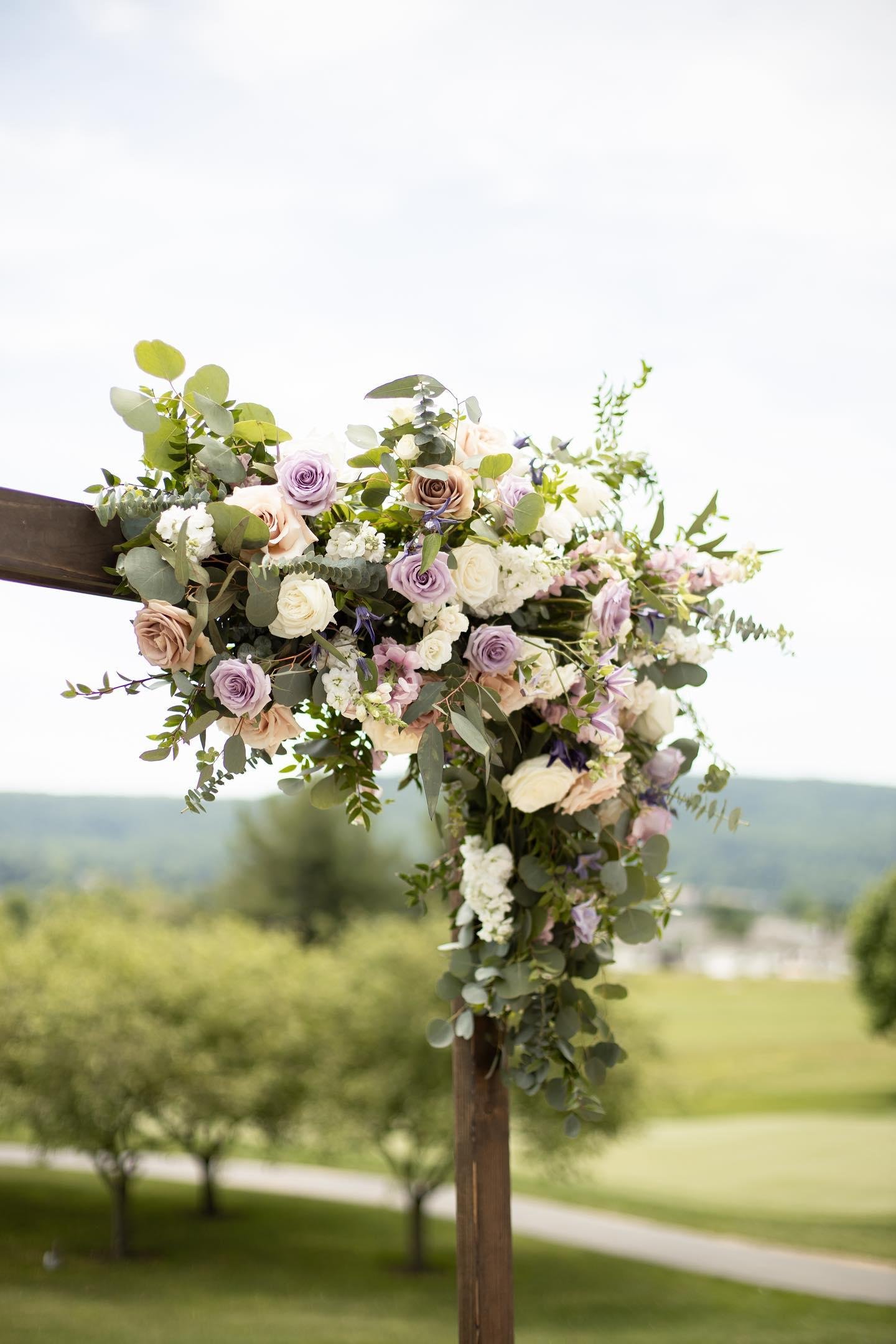 A floral arrangement with white, purple, and tan roses along with greenery on a wooden structure at an outdoor wedding venue.