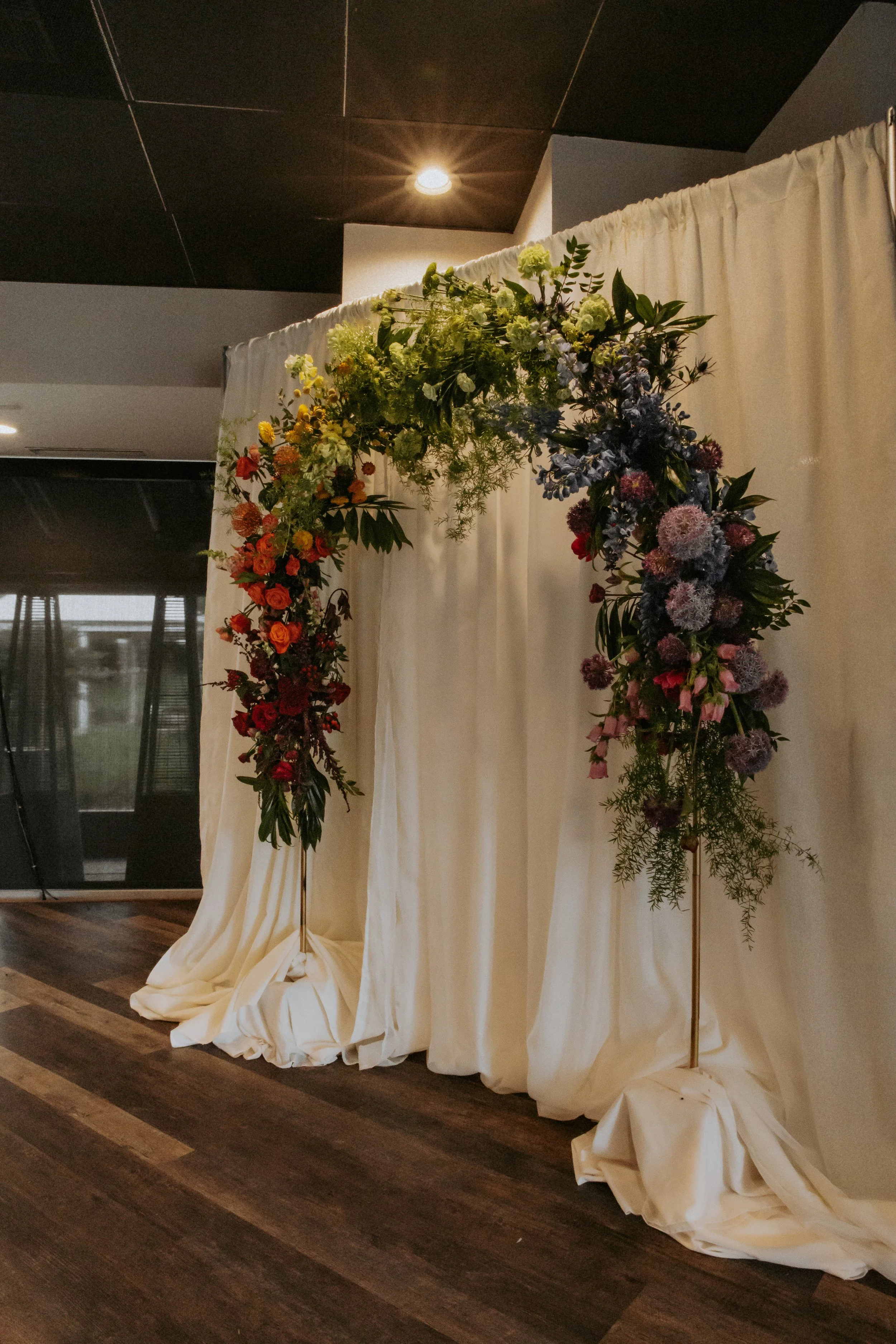 Floral arch with mixed flowers in red, white, and purple, set against white drapery background for a wedding or event.