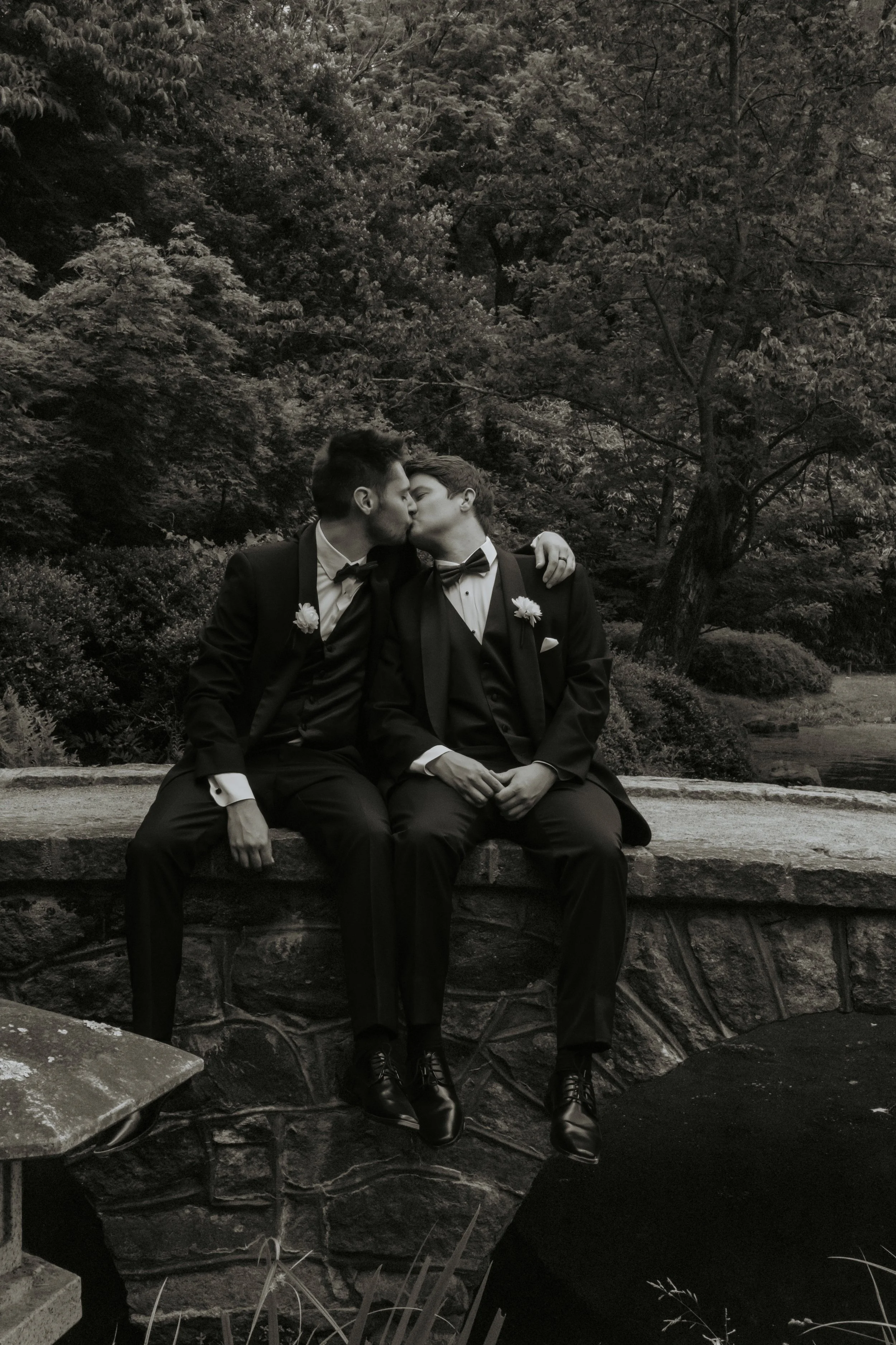 Two men in tuxedos kissing on a stone ledge in a natural outdoor setting.