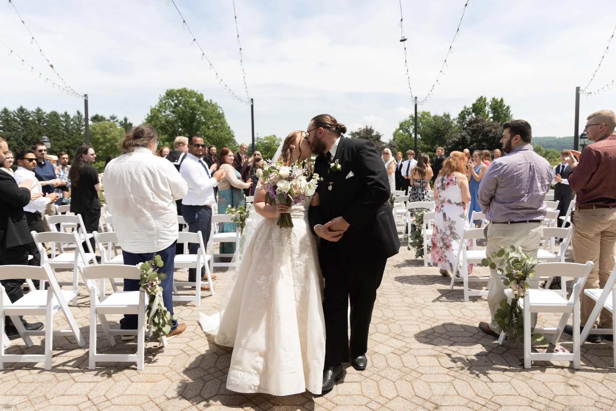 A wedding couple, the bride in a white dress holding a bouquet and the groom in a black suit, kiss under a string of outdoor lights at a wedding ceremony with guests seated around them, greenery in the background.
