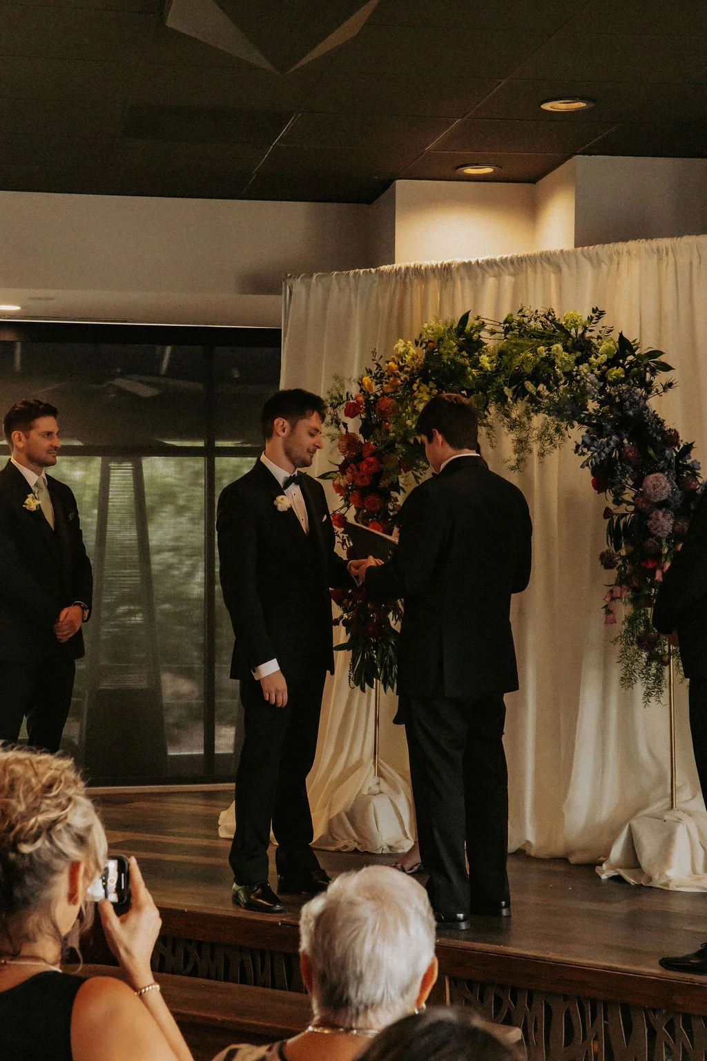 A wedding ceremony taking place indoors with three men in tuxedos, one of whom is reading from a book while facing a floral arch decorated with colorful flowers. Guests are seated and some are taking photos.