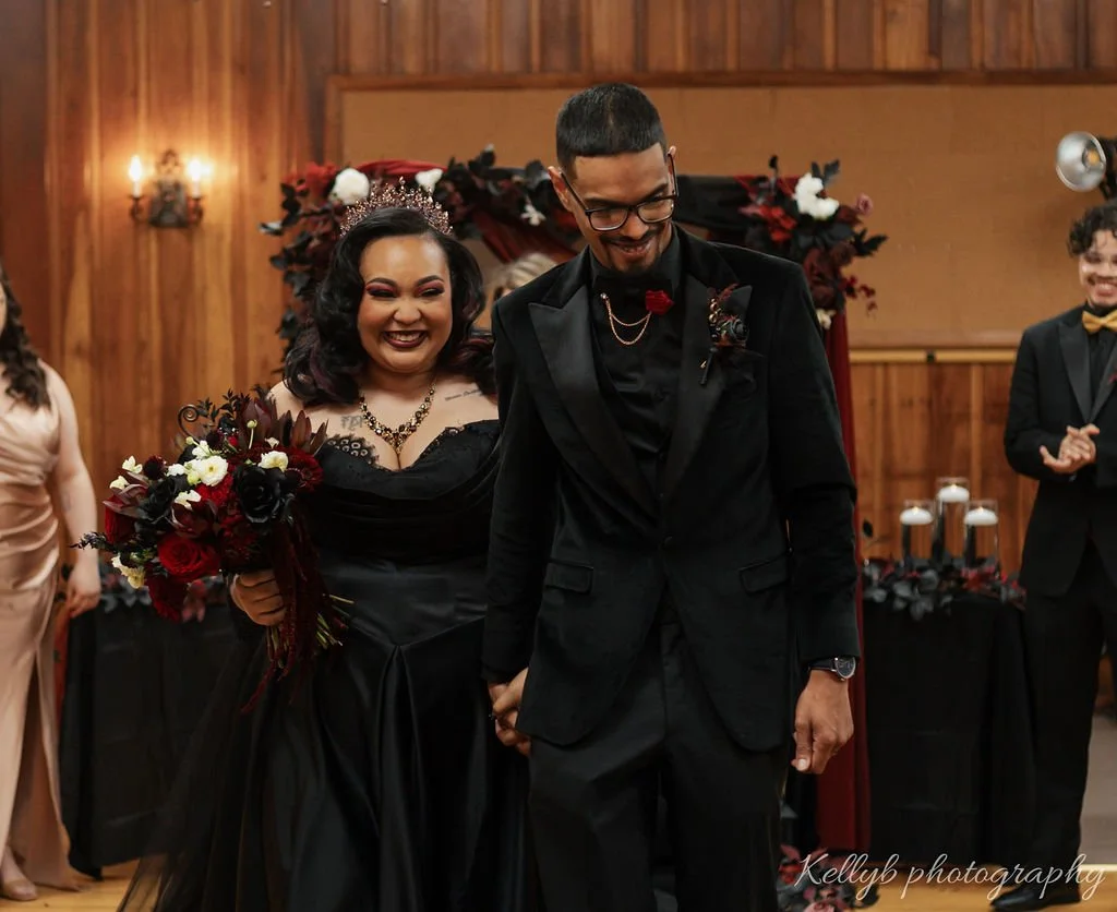 A joyful couple at a wedding ceremony, with the woman wearing a black dress and crown, holding a bouquet of dark and white flowers, and the man in a black tuxedo with glasses, holding the woman's hand. They are surrounded by smiling guests in formal 