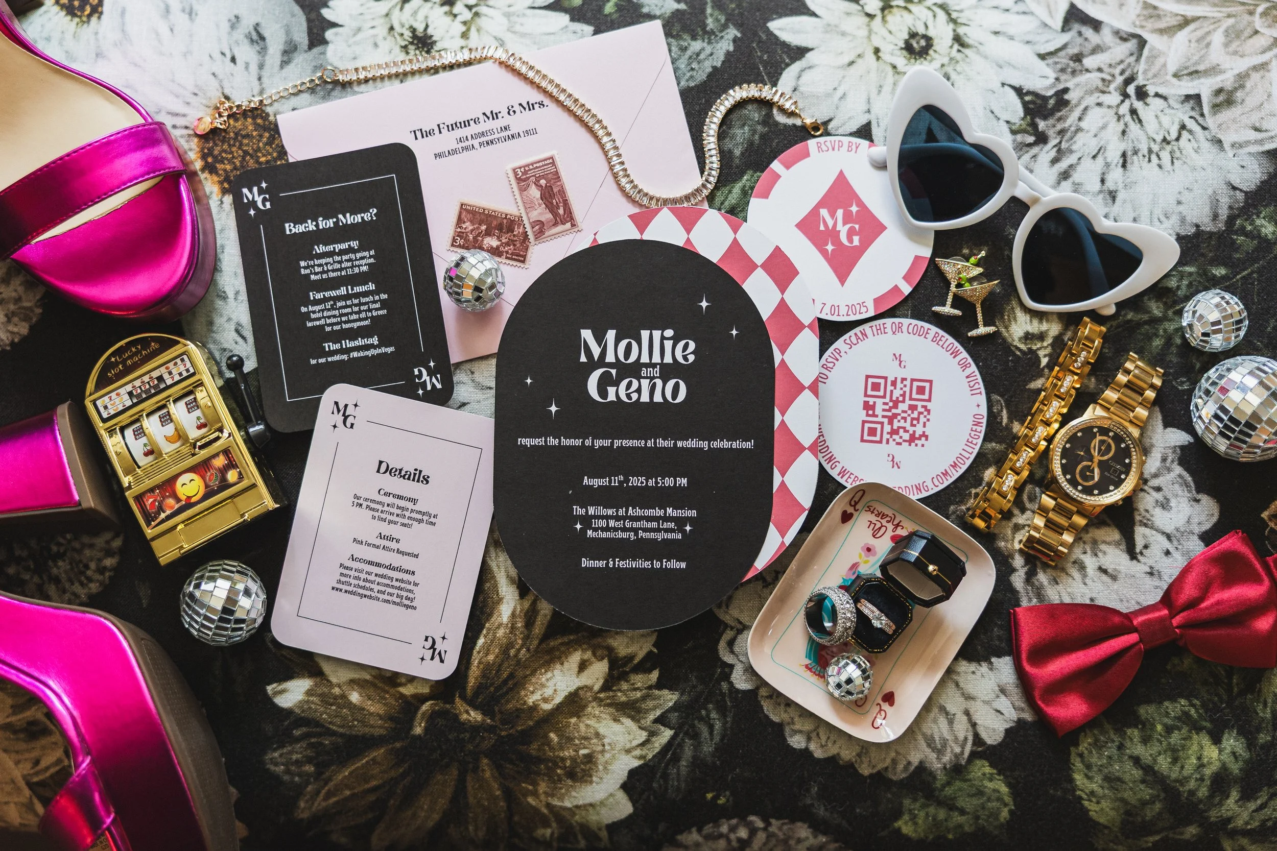 Wedding invitation, jewelry, rings, sunglasses, disco balls, a watch, a pink bow, a slot machine, a pink high heel shoe, and various party accessories arranged on a floral tablecloth.