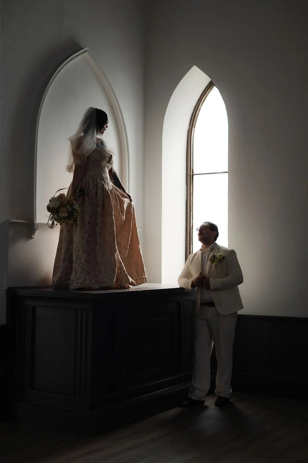 A bride on a raised platform holding a bouquet stands near a groom, who is looking up at her, inside a church with a tall arched window backlighting them.