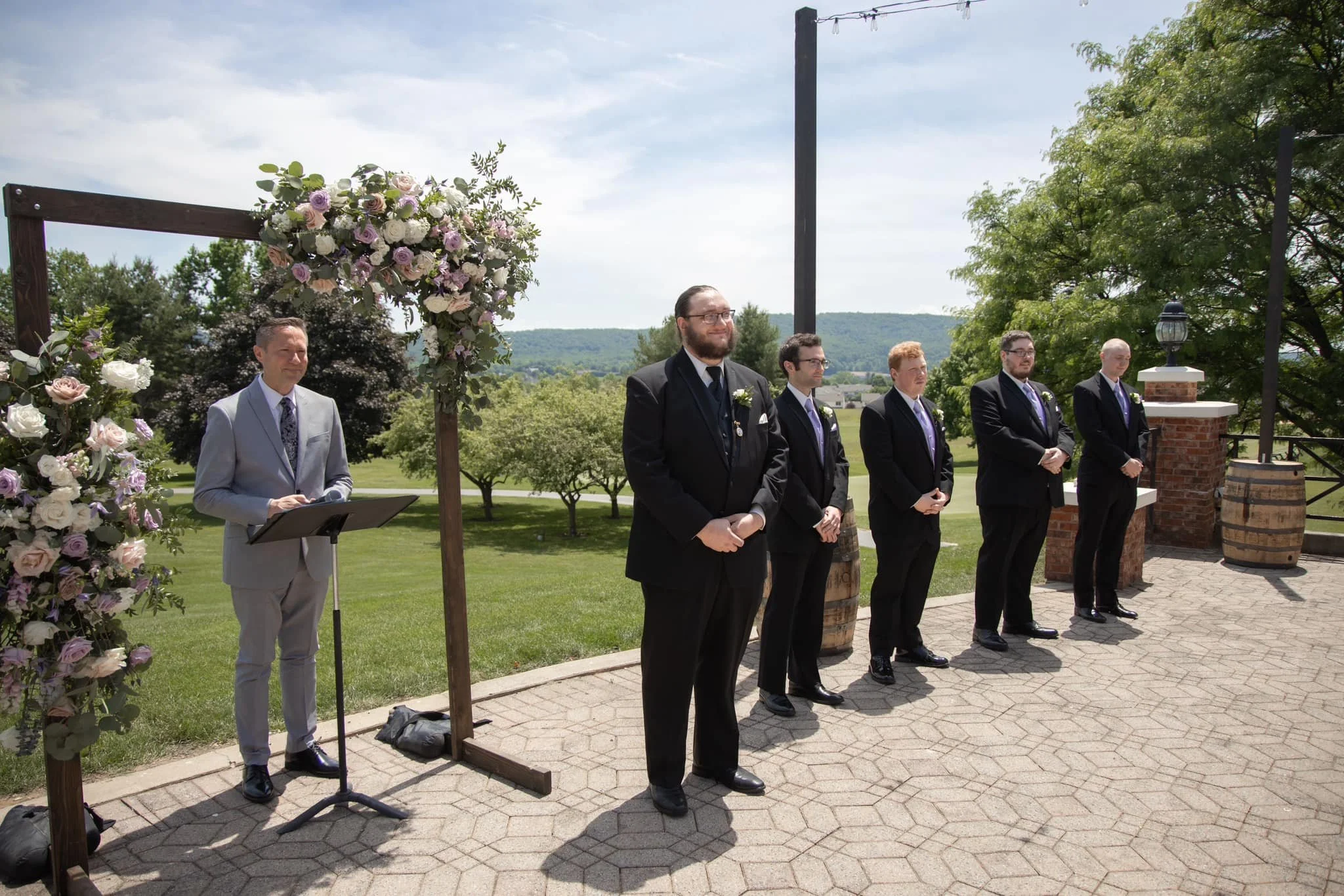 A wedding ceremony outdoors on a sunny day with six men dressed in formal suits standing on a brick patio, facing forward. There is a floral arch with pink and white flowers to the left, and scenic trees and hills in the background. One man, in a lig